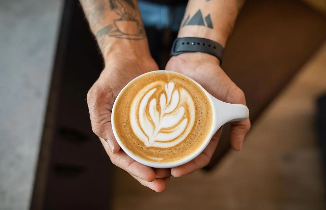 Person holding a white ceramic cup of latte with latte art on top, showing a leaf or tulip design.