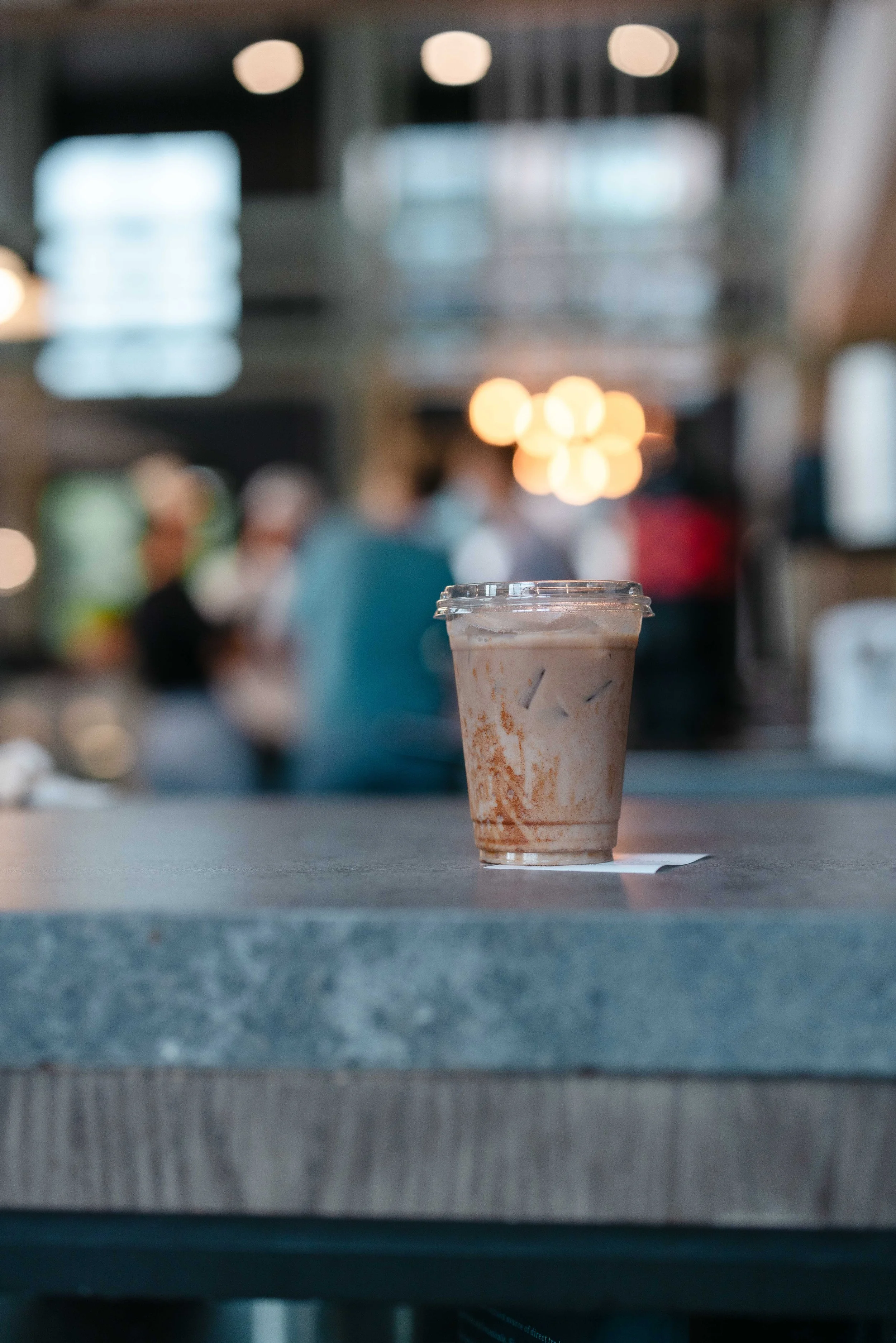 A plastic cup of iced coffee or chocolate frappuccino on a table in a busy coffee shop with blurred customers in the background.