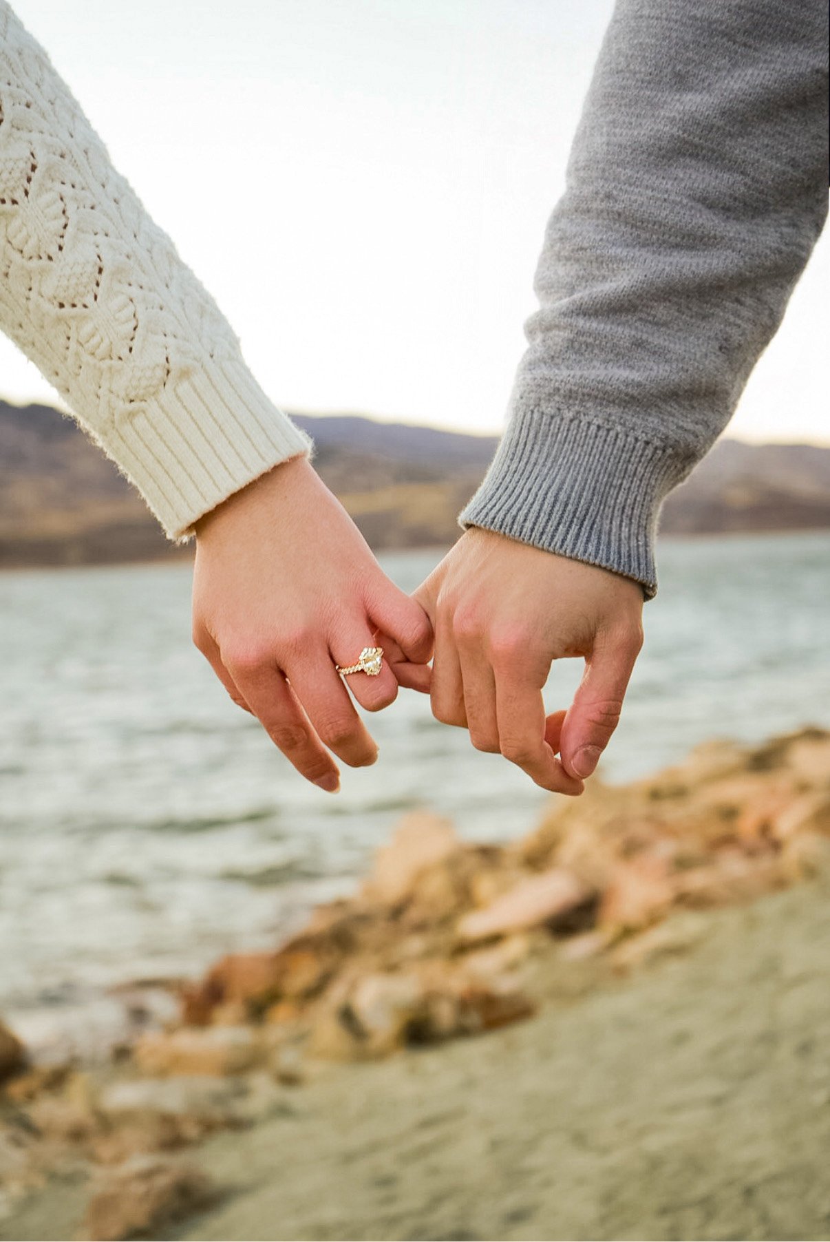 Close-up of two people holding hands with a background of water and mountains, one person wearing a white knitted sweater and the other a gray sweater, with a diamond engagement ring on the left person's finger.