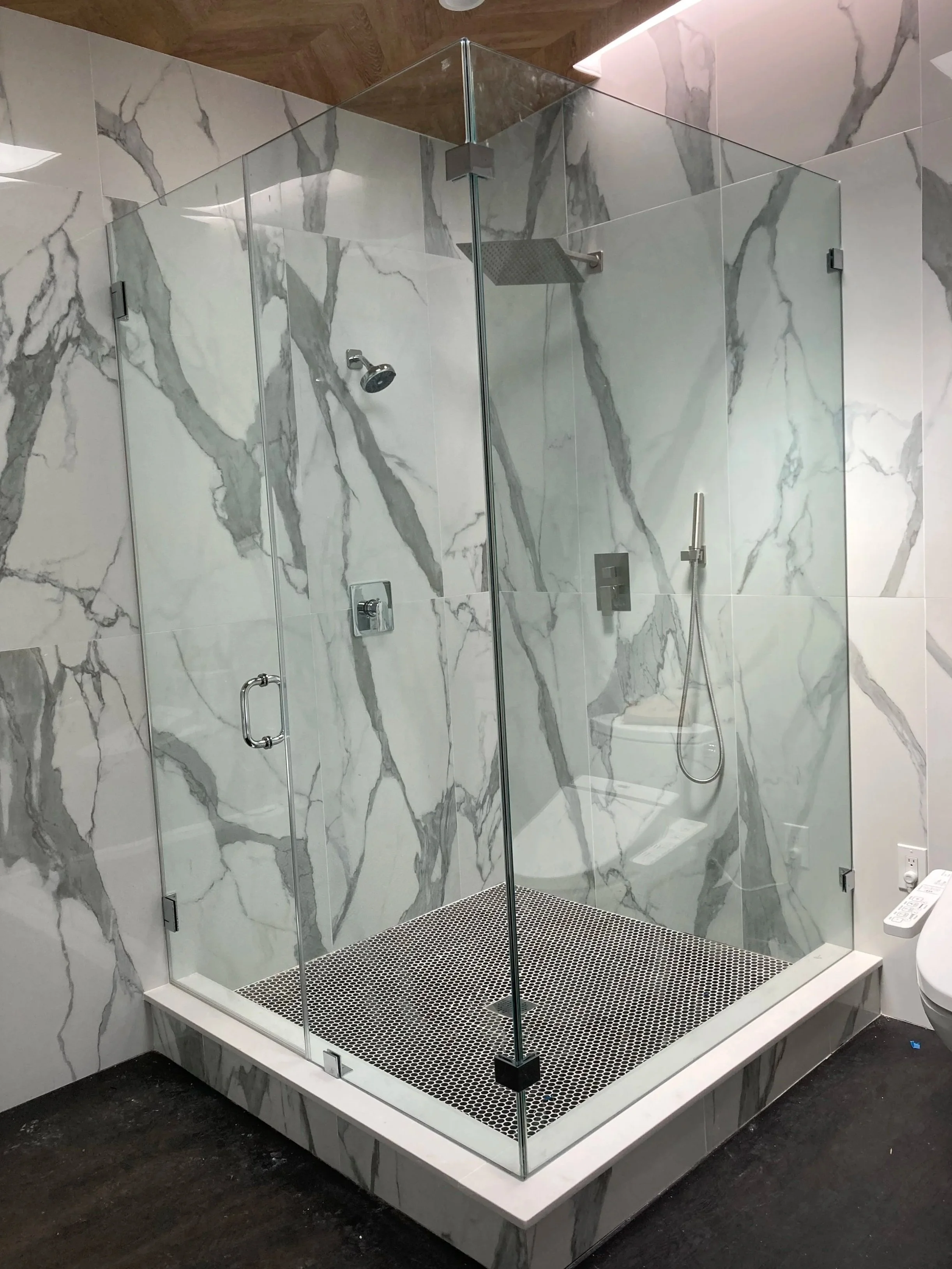 A modern glass shower with marble walls, a rainfall showerhead, a handheld shower, and a black and white tiled floor, adjacent to a toilet.