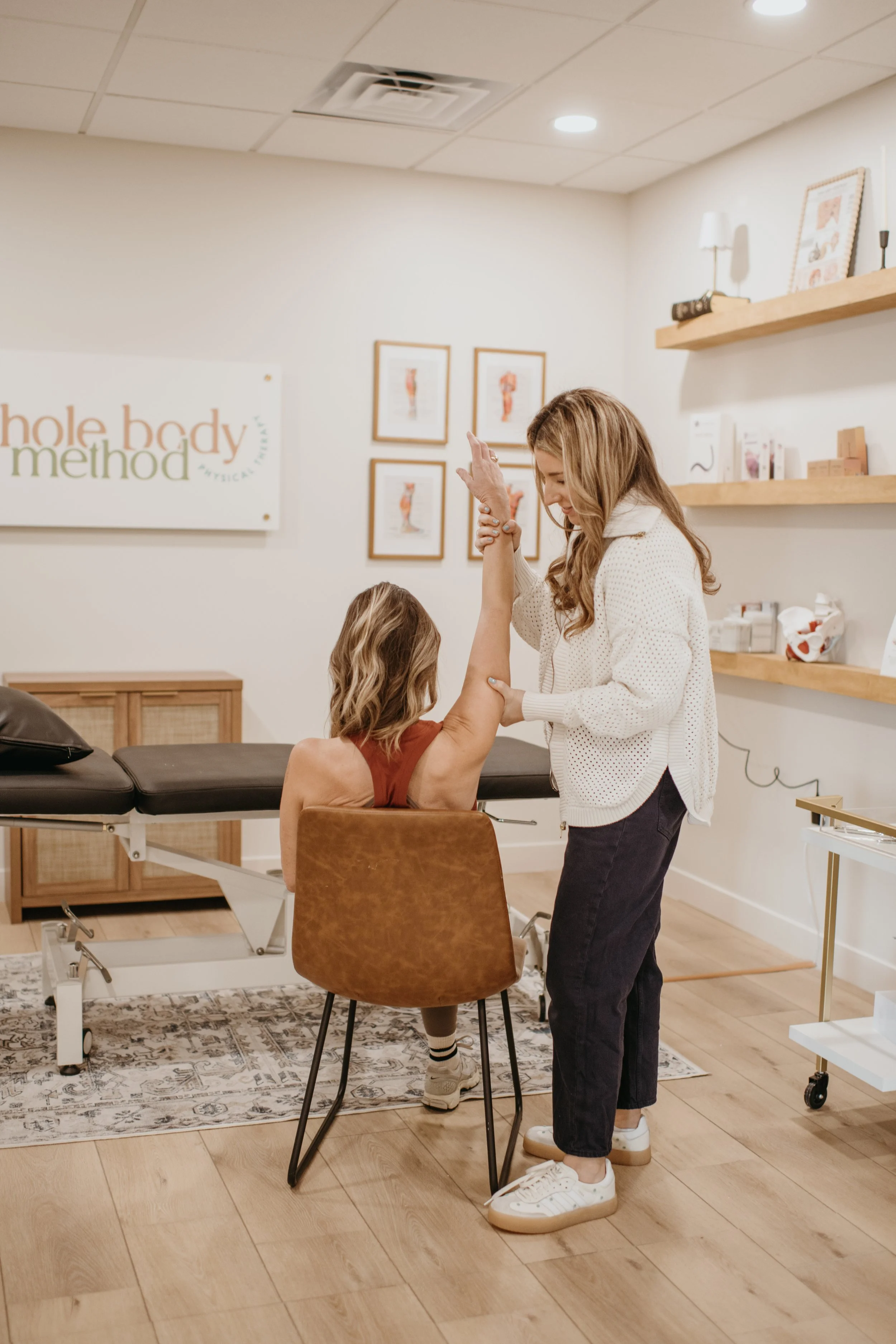 A woman receiving physical therapy from a therapist in a clinic. The woman is seated with her arm raised, and the therapist is holding her arm and smiling.