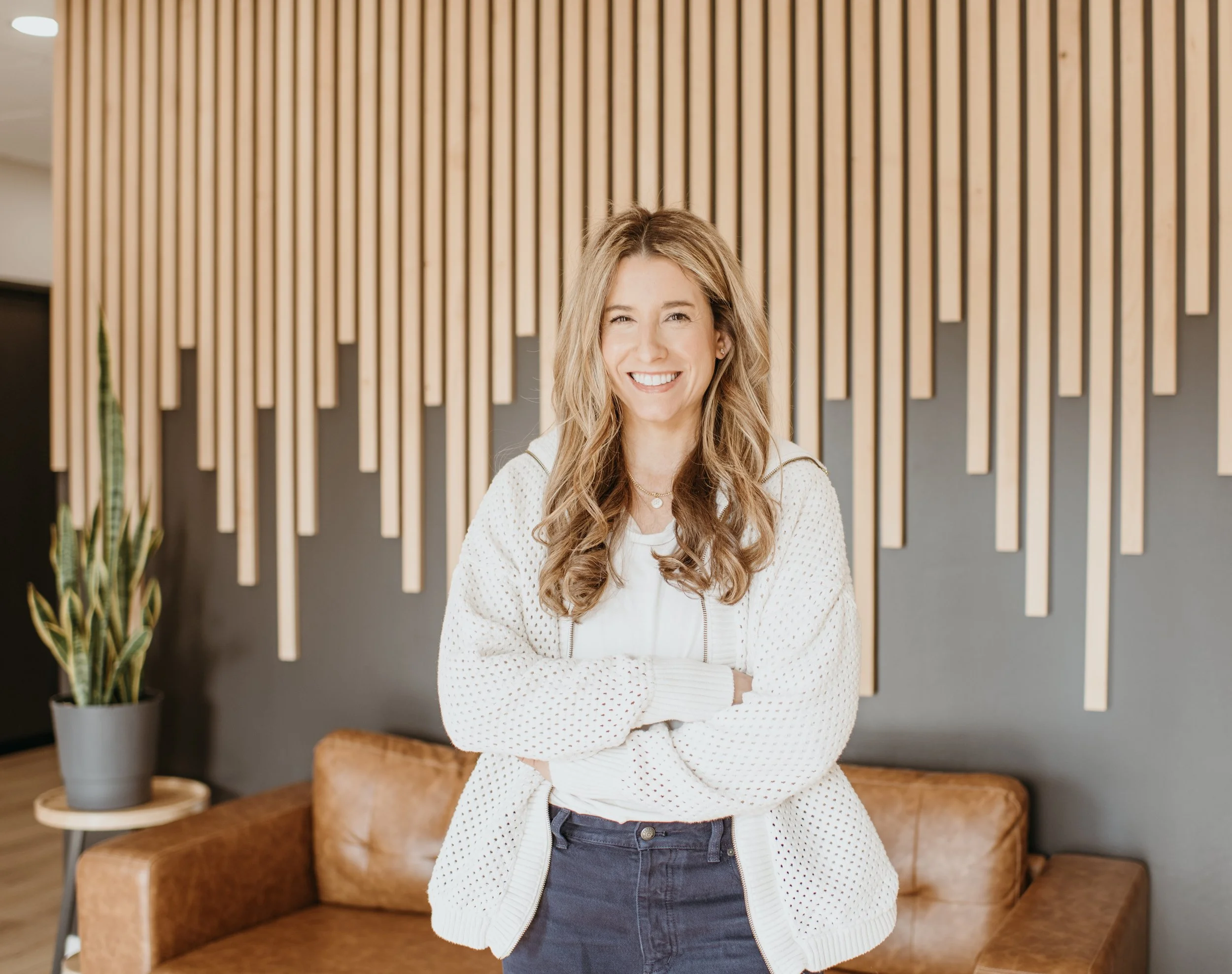 A woman with long wavy hair smiling with arms crossed, standing in front of a modern wooden wall with a potted plant nearby.