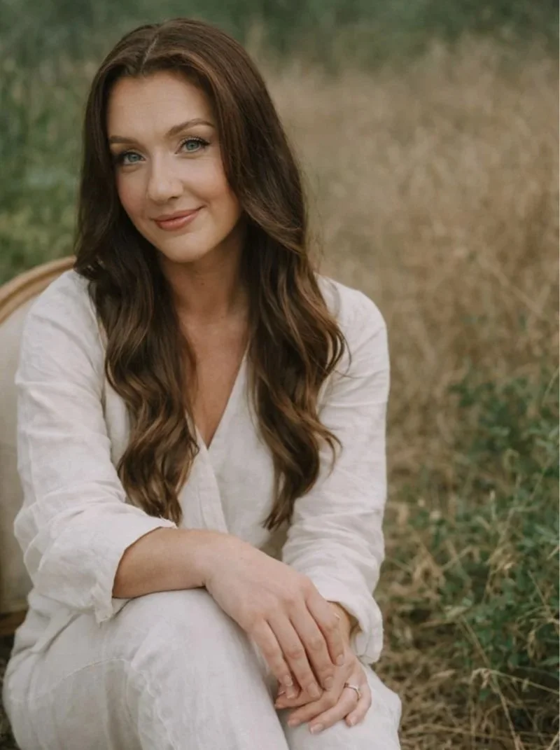 A woman with long, wavy brown hair and blue eyes sitting outdoors in a natural setting, wearing a cream-colored linen outfit, smiling softly.