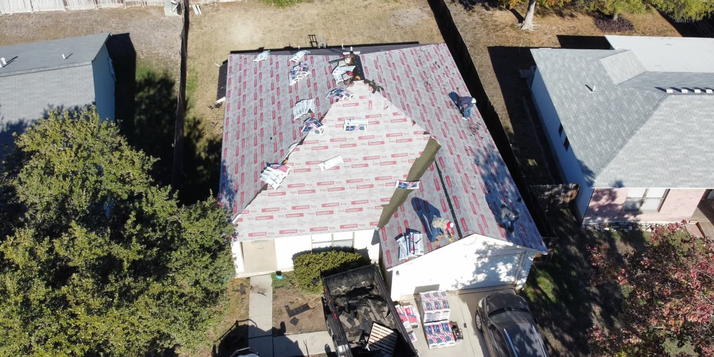 Aerial view of a house under construction with a partially installed roof. Building materials and tools are scattered on the roof. Nearby are neighboring houses and trees.