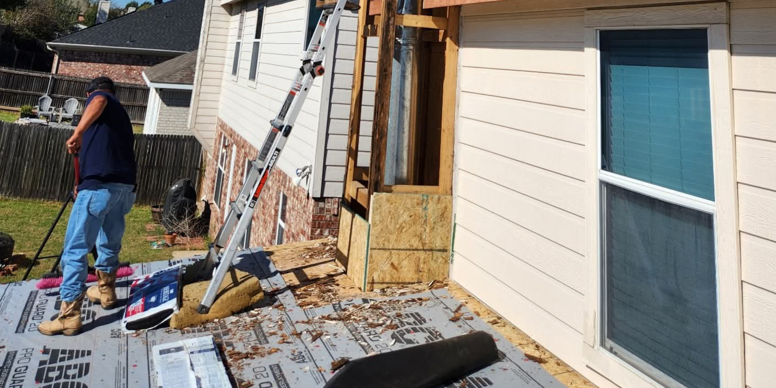 Construction worker standing outdoors next to a house, working on an extension or renovation. The house has white siding, and construction materials and tools are around. A ladder is leaning against the house. The scene takes place in a backyard with a fence and neighboring houses visible.