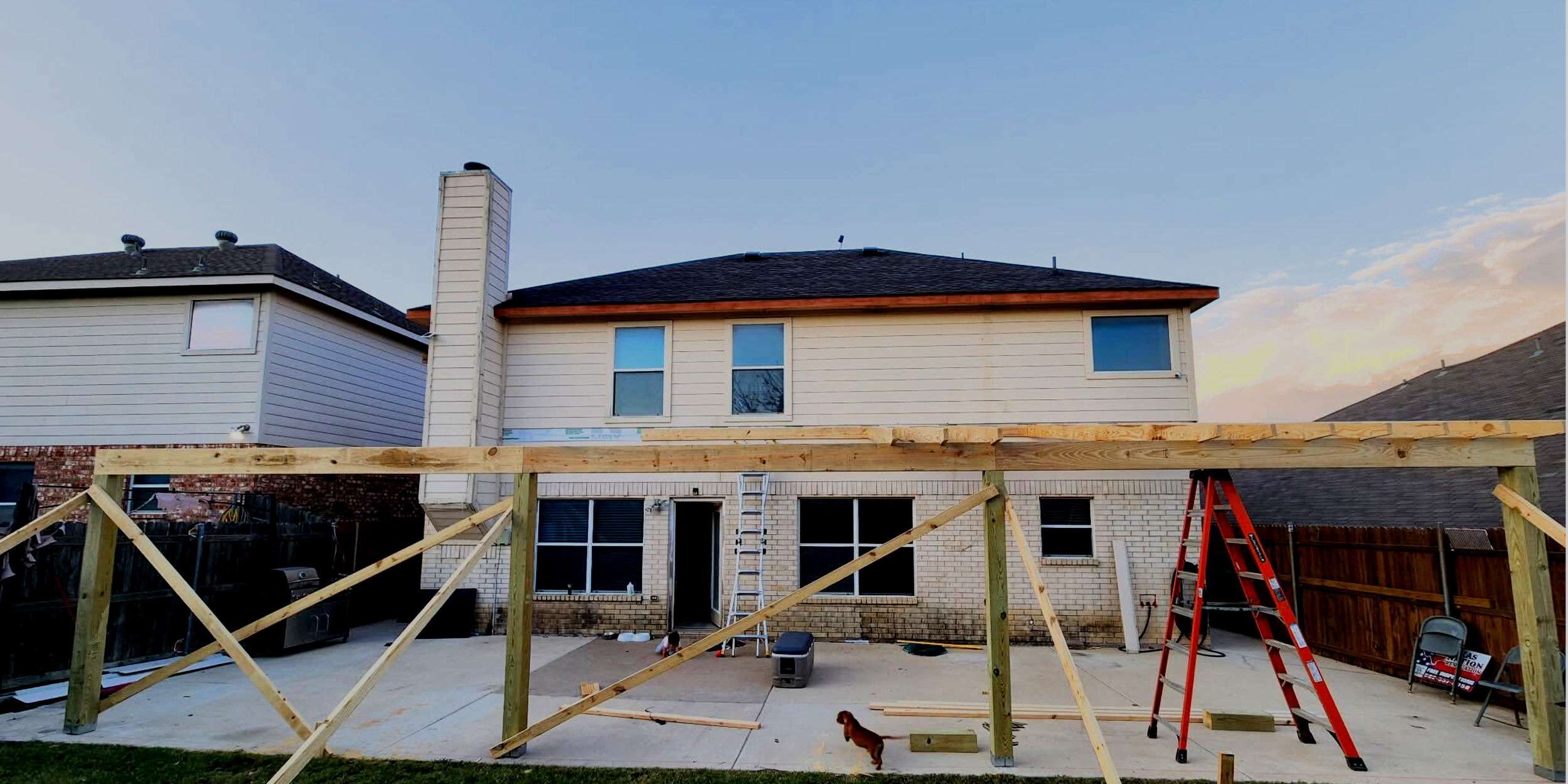 Backyard construction site with wooden framework, ladders, tools, and a small dog, in front of a two-story house with brick and siding exterior during sunset.