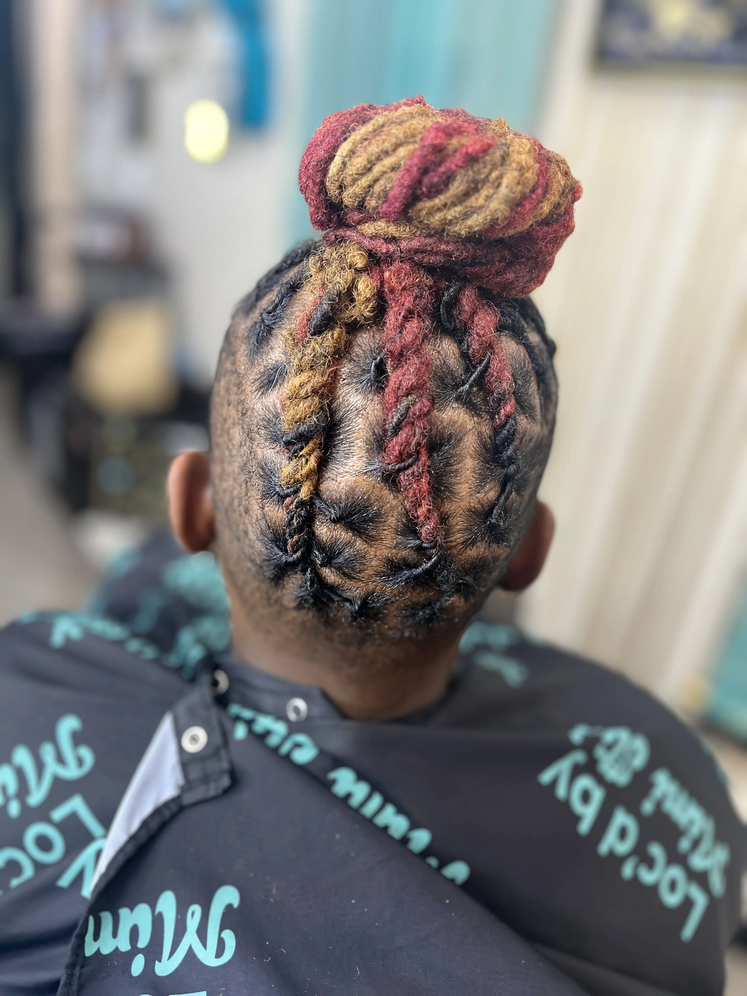 Back of a person's head with dreadlocks styled into a bun, with some dreadlocks dyed red and blond, sitting in a barber chair.
