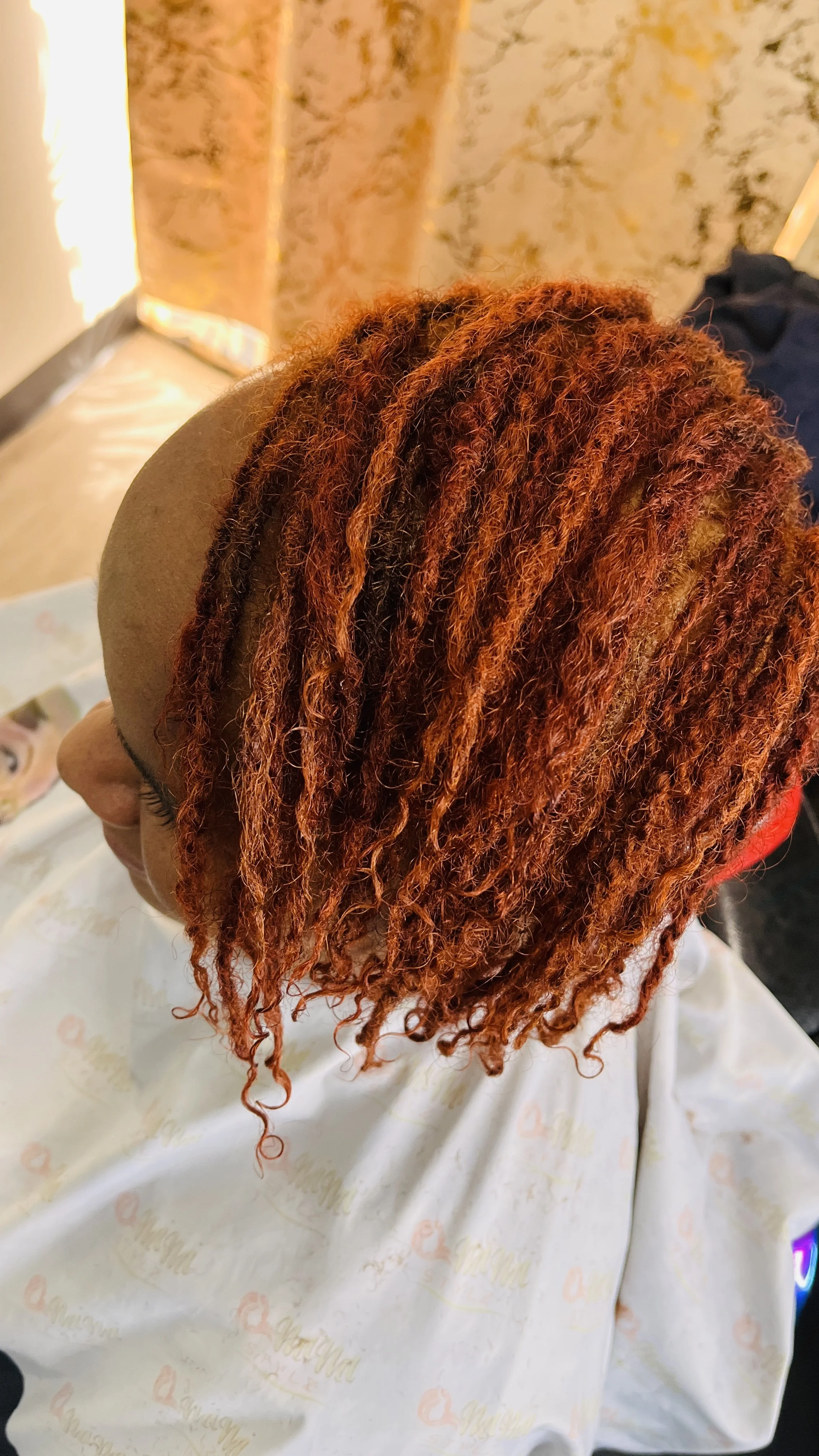 Person with reddish-orange dreadlocks sitting in a salon chair, wearing a beige hat and a salon cape with faint logo prints.