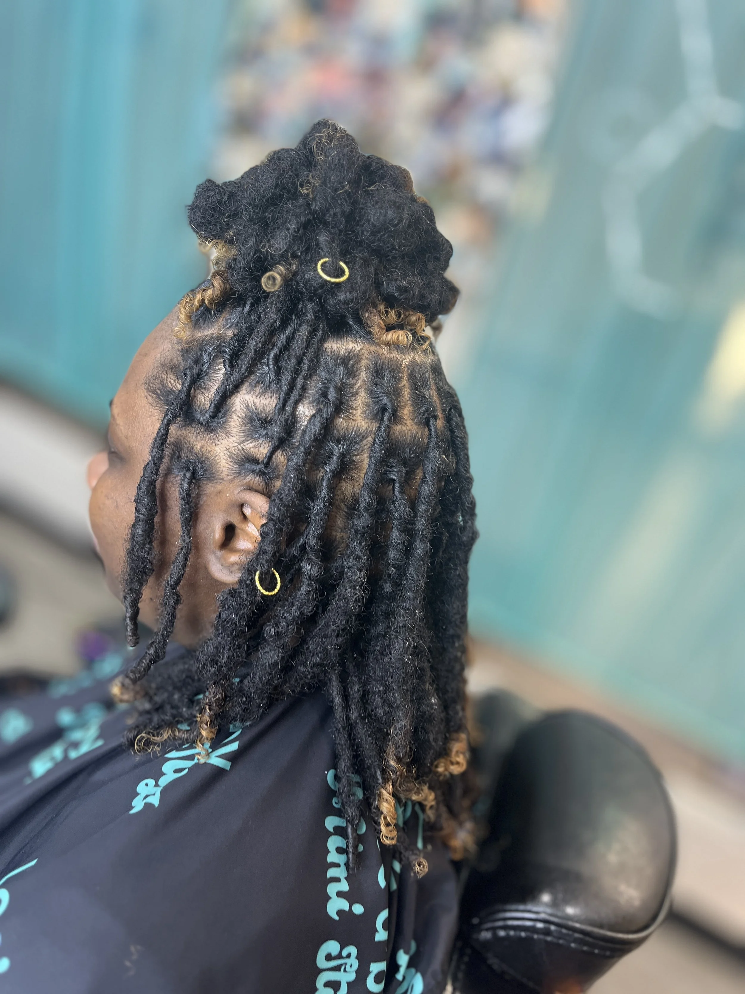 A person with dark skin and thick black and brown dreadlocks, wearing gold hoop earrings, seated on a black chair with a textured surface, background blurred with teal-colored walls.