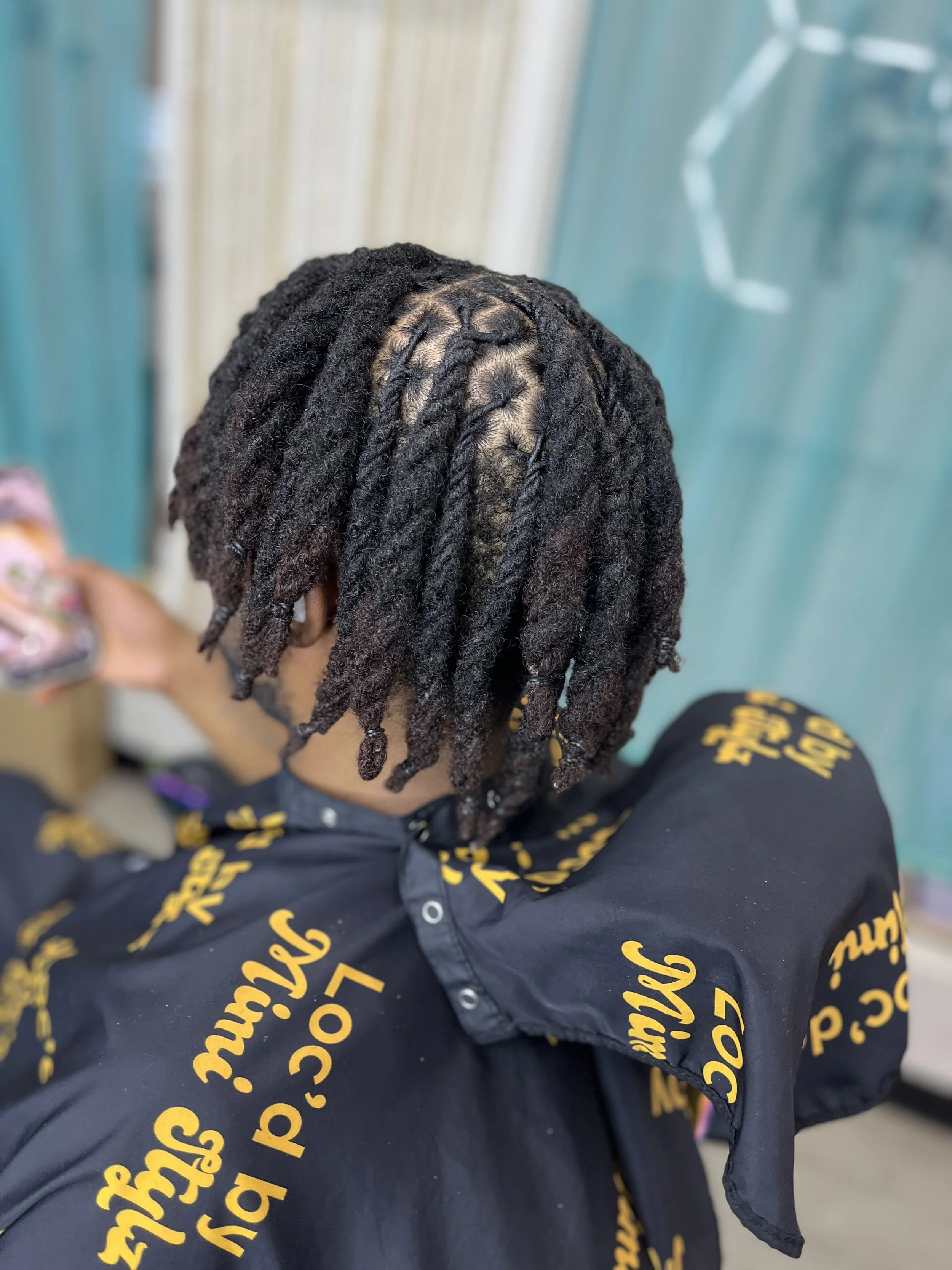 A person with short dreadlocks sitting down, wearing a black jacket with yellow writing, in front of a teal background.