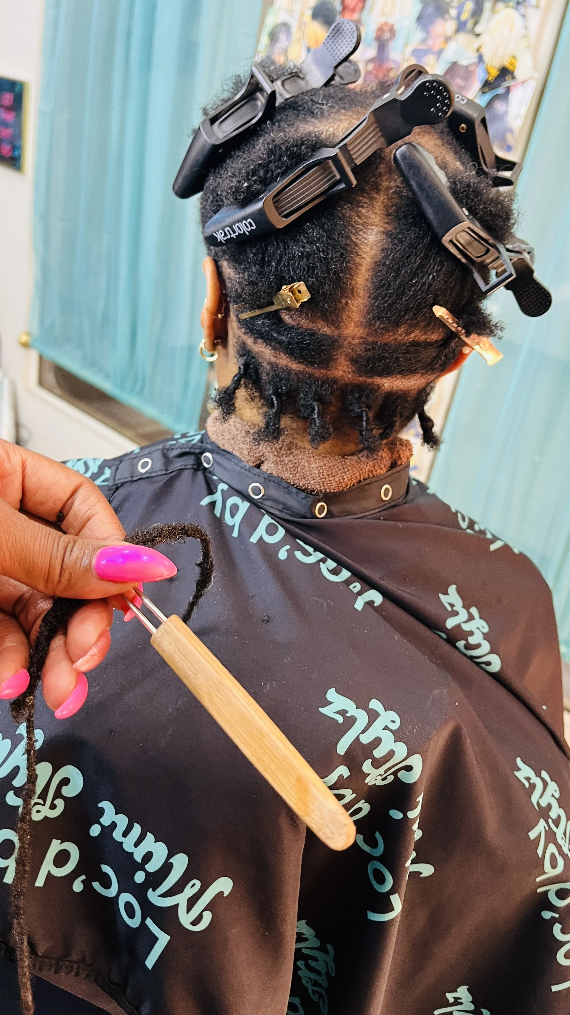 A woman with short, natural hair getting her hair styled at a salon. She is wearing a black cape with green writing and has multiple hair clips and rollers securing her hair.