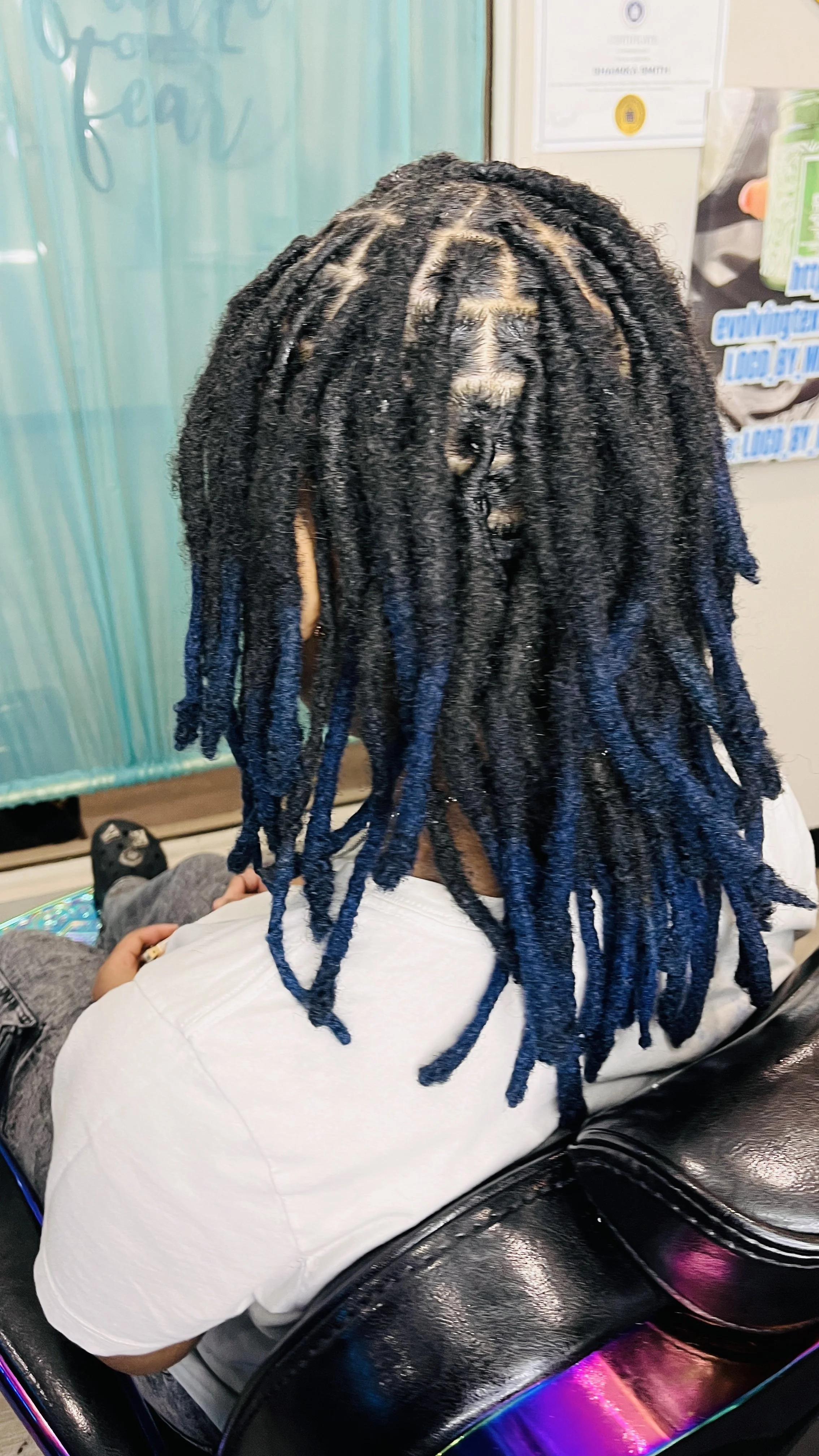 Back view of a person with blue dreadlocks, wearing a white shirt, sitting on a black chair with iridescent accents in an indoor setting.