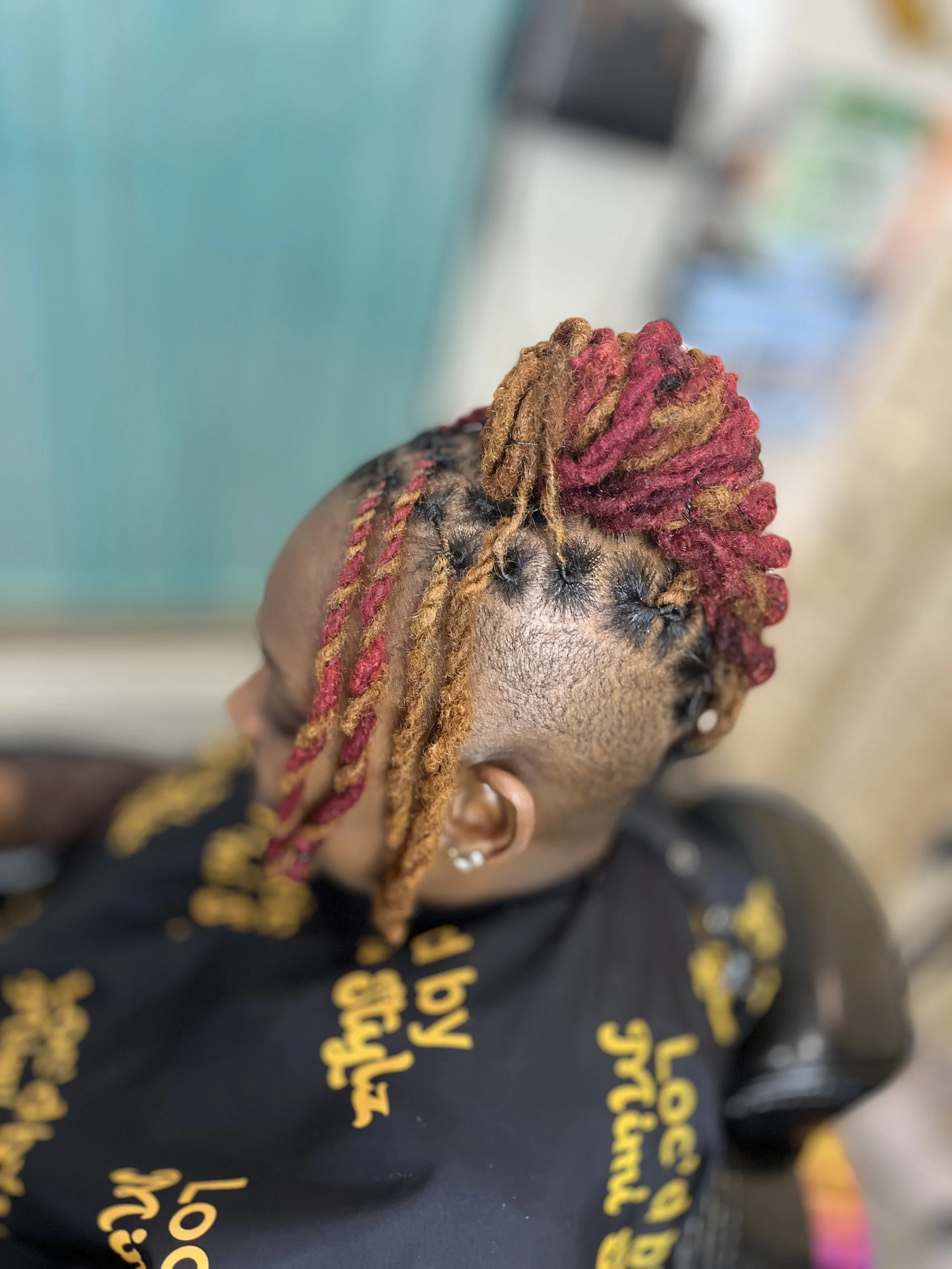 A person with braided and colored hair looking down with a blurred background. The person is wearing a black shirt with yellow text.