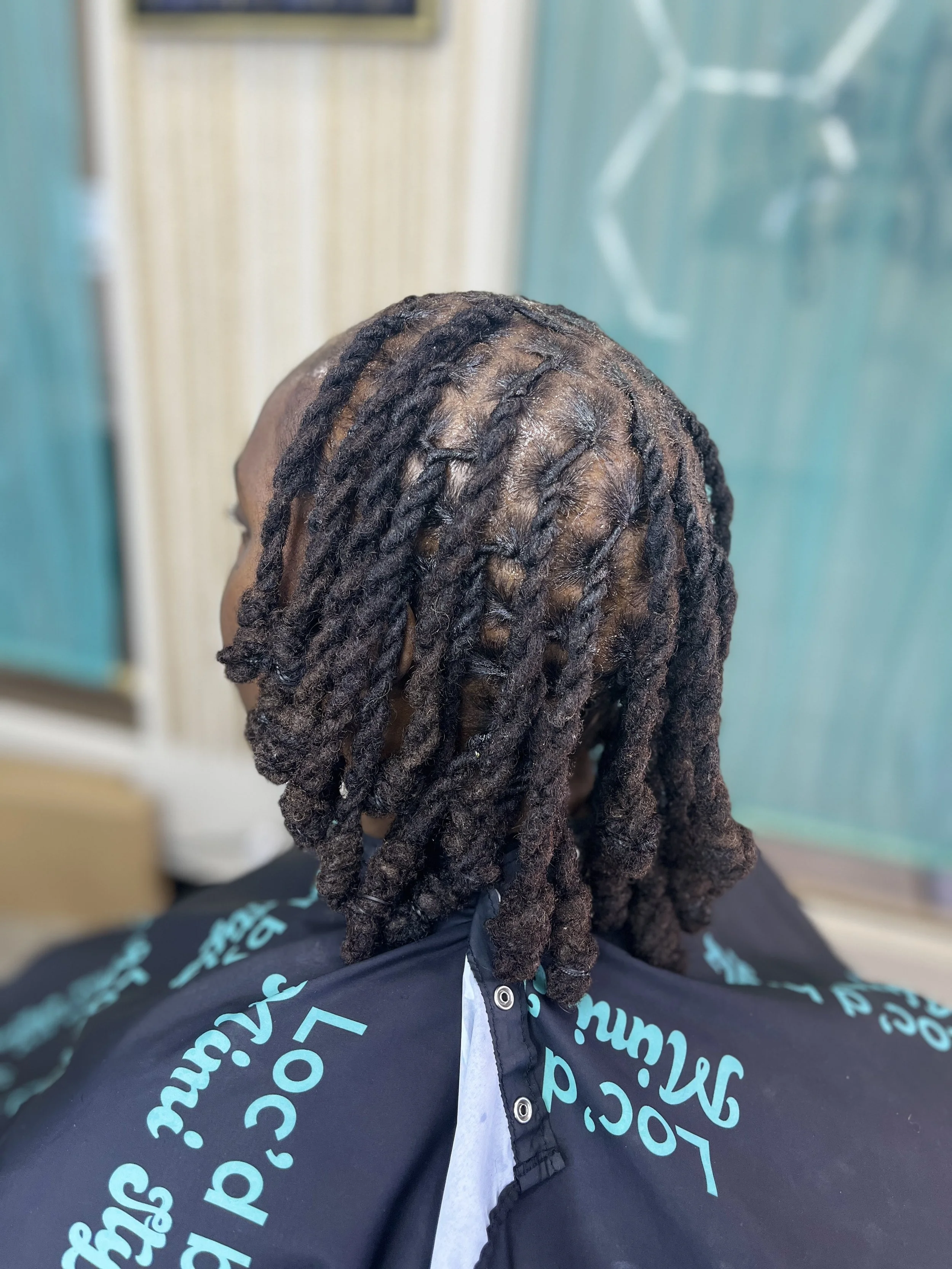 The image shows a person with dark brown dreadlocks wearing a black jacket with blue text on it. The background has a blurred blue wall with a window or glass panel.