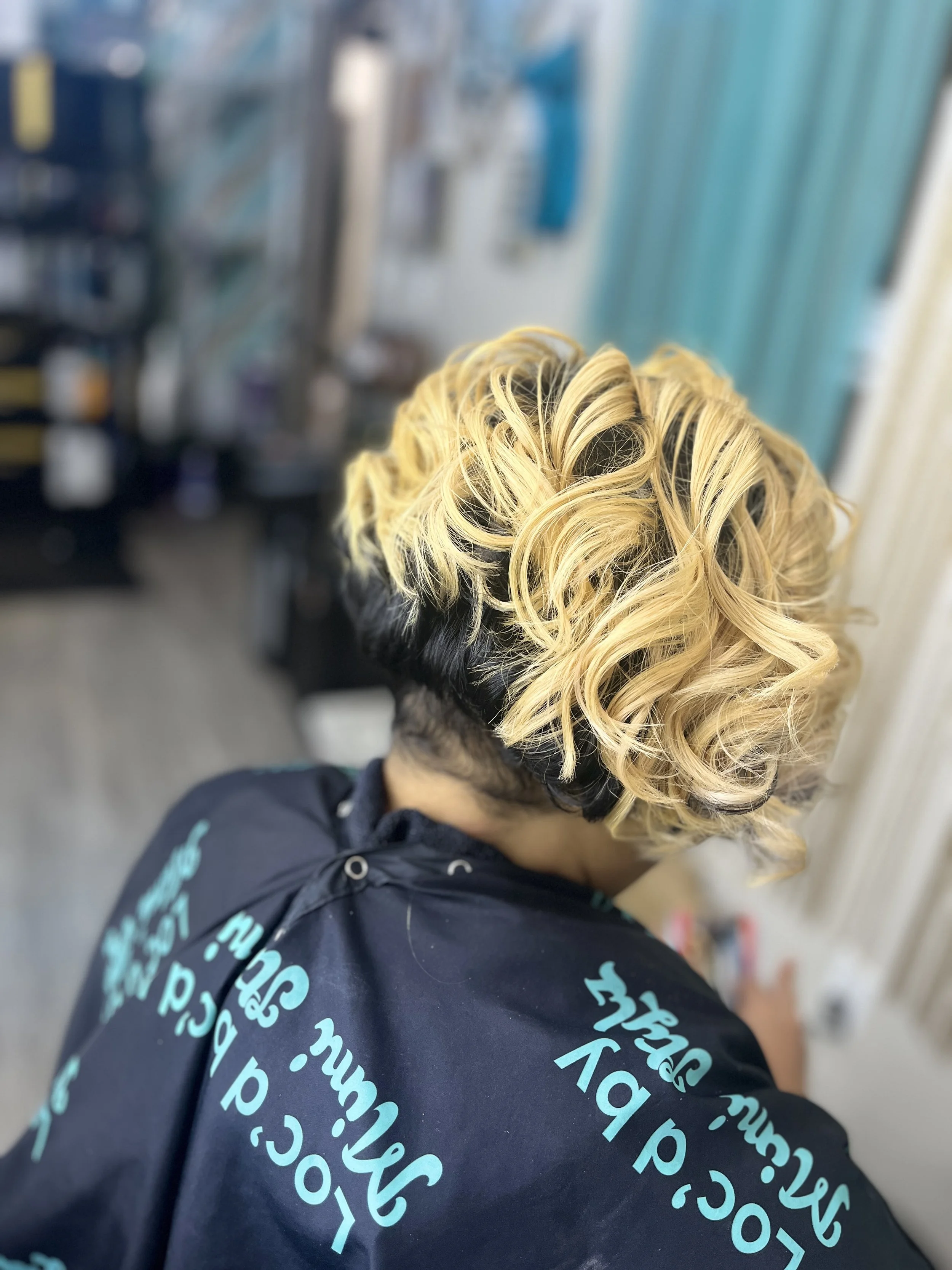 Top view of a person's head with blond curly hair and black roots, wearing a black hoodie with blue text, in a setting with industrial and blue-green background elements.
