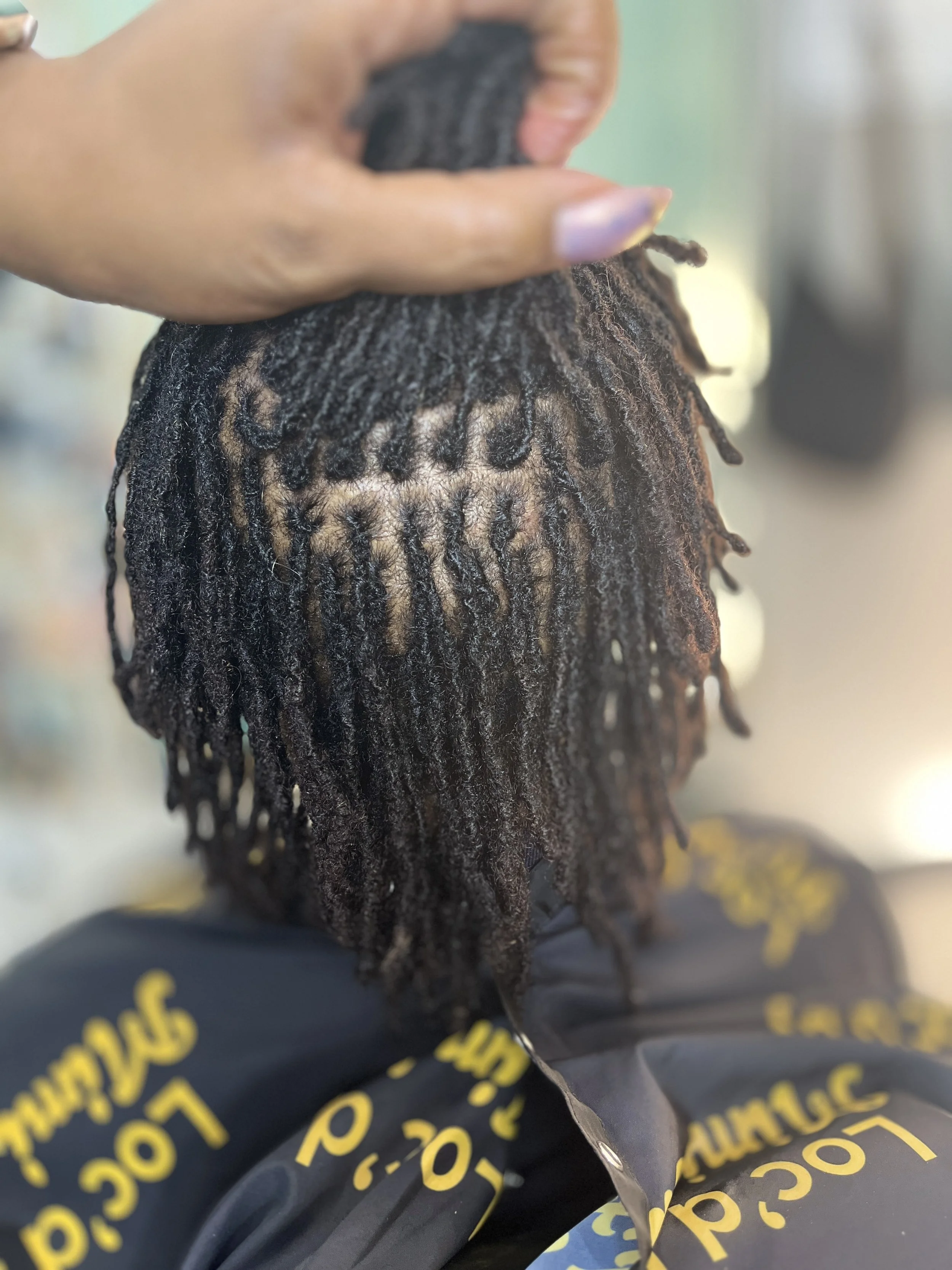 Close-up of someone holding a bundle of dreadlocks hair, with a blurred background and some yellow text on dark clothing visible at the bottom.