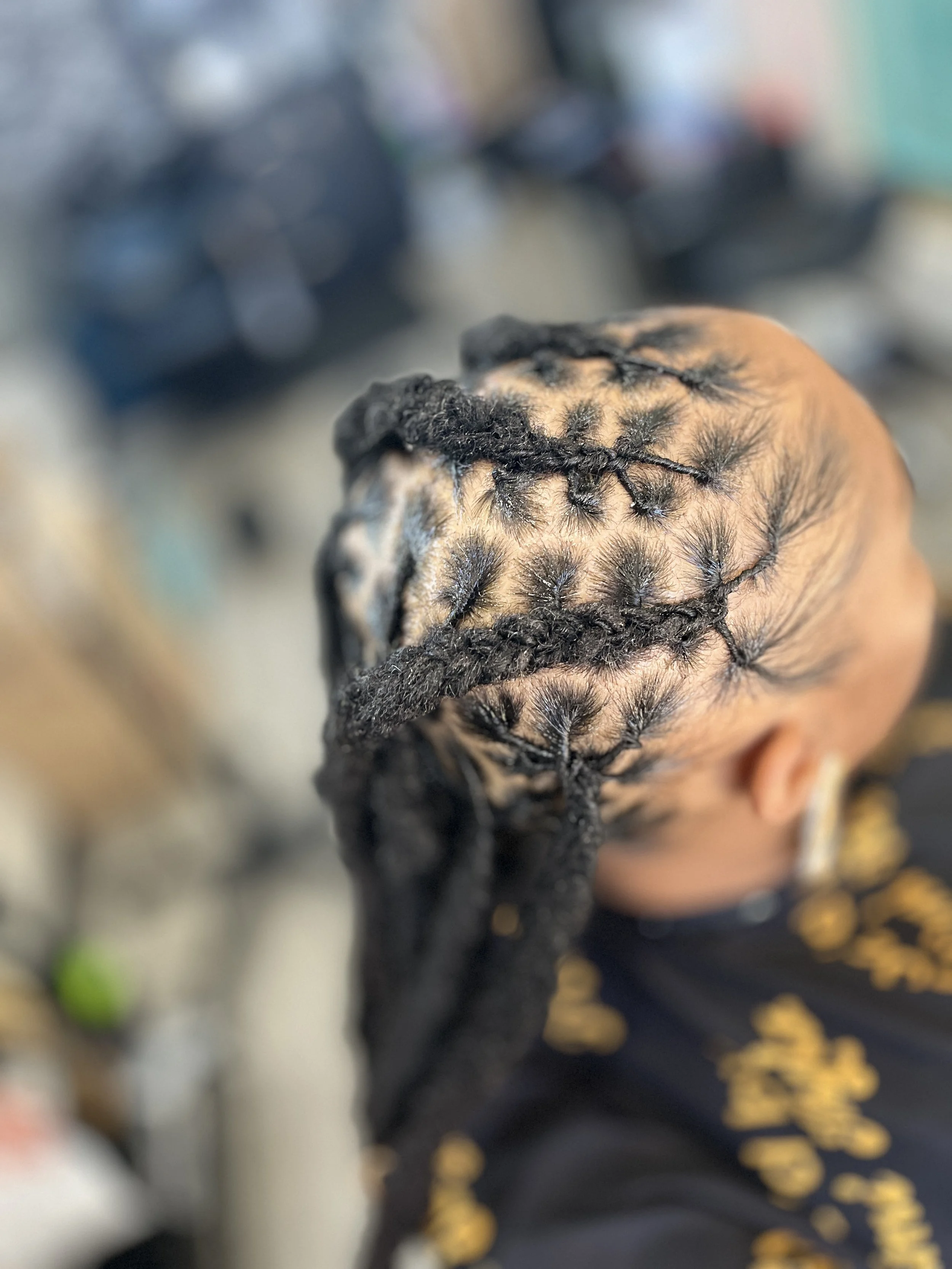 A person with braided hair, viewed from above, with a blurred background including a person wearing a black shirt with gold print.
