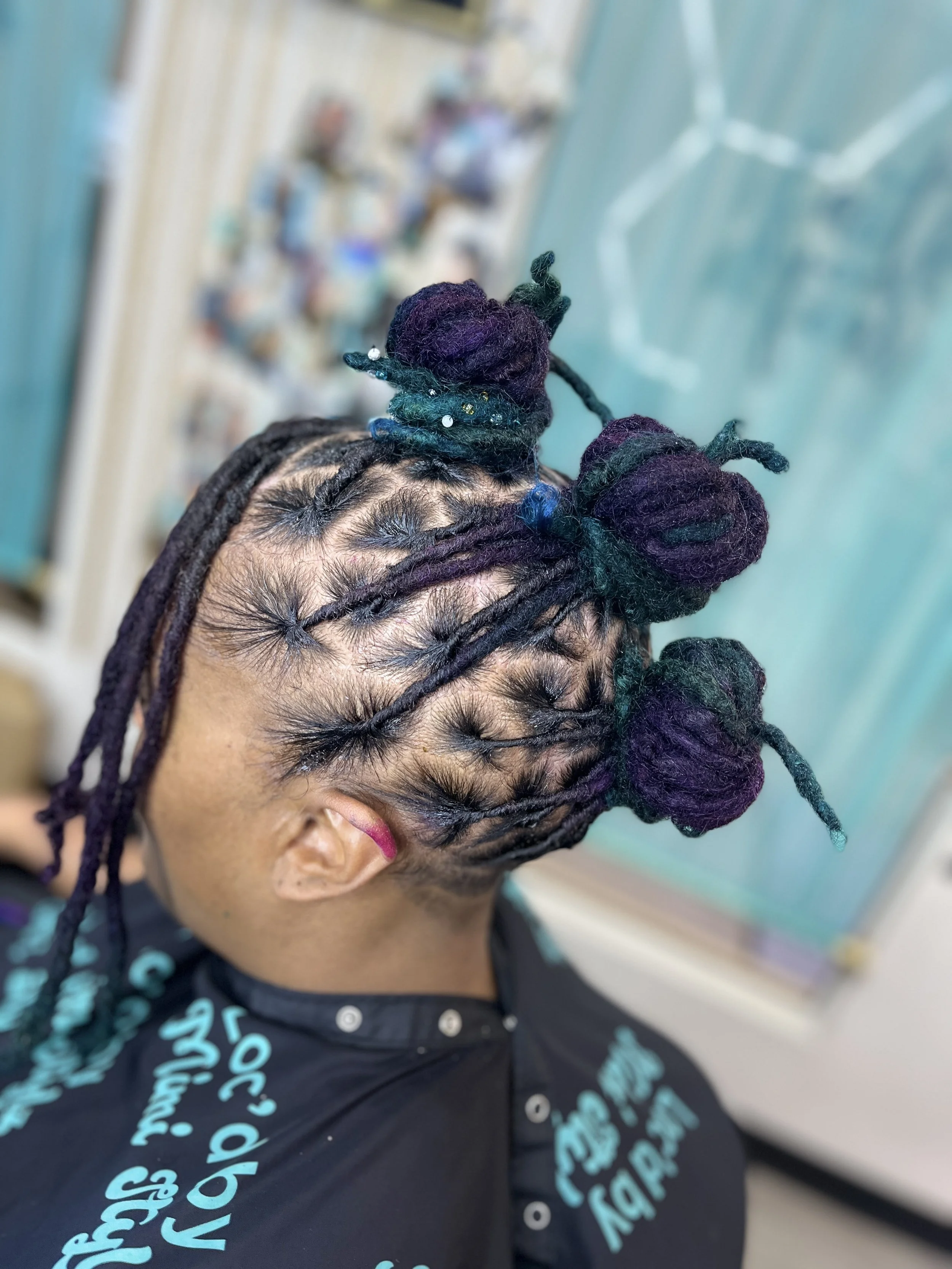Close-up of a person's head with black dreadlocks styled into buns, with some beads and small decorations, and a purple hair accessory. The person is wearing a black shirt with turquoise text. The background shows a blurred room with various items.