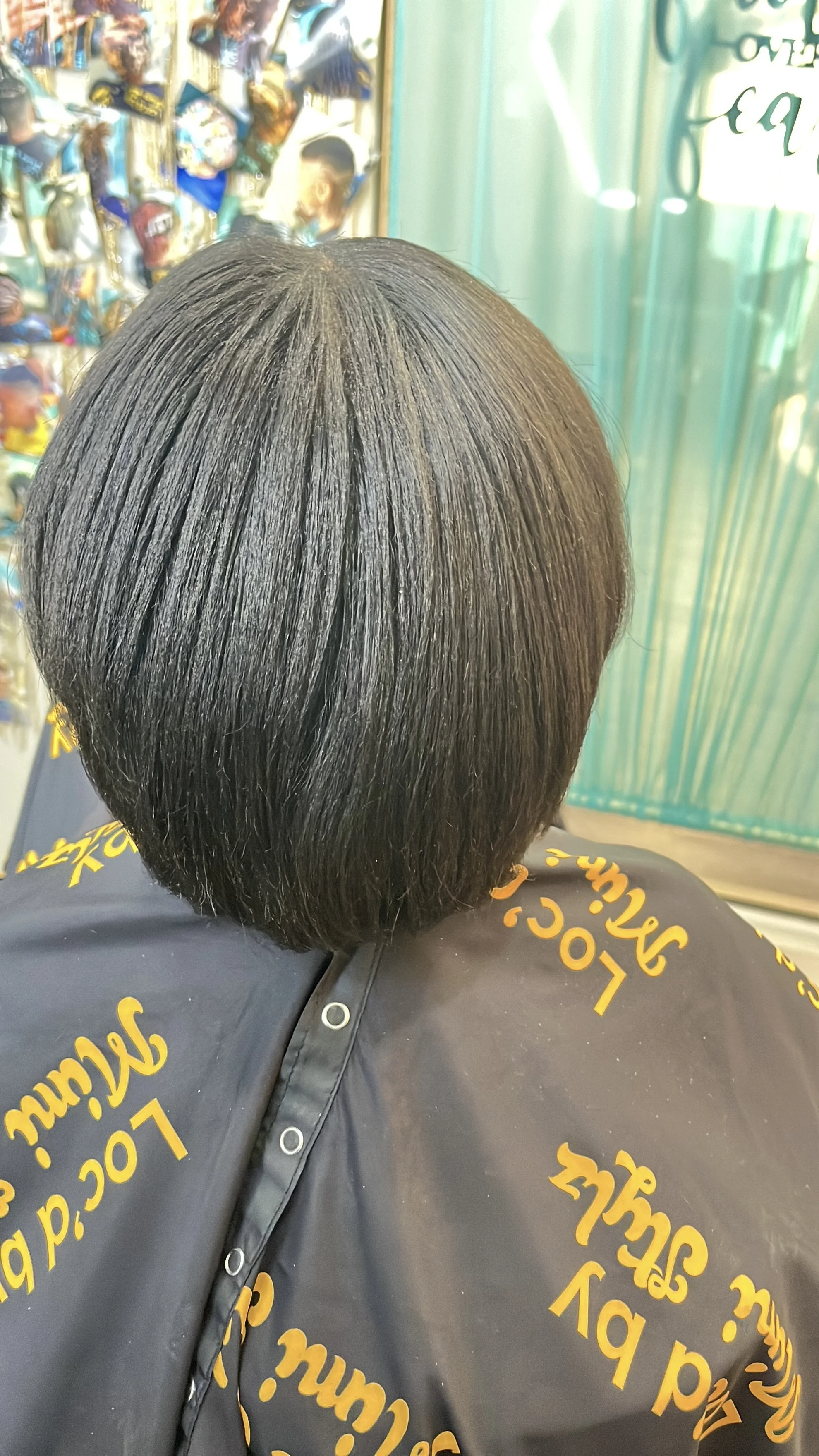 The image shows the back of a person's head with a neat, short, shoulder-length black haircut, sitting in a salon with a black cape that has yellow text on it.