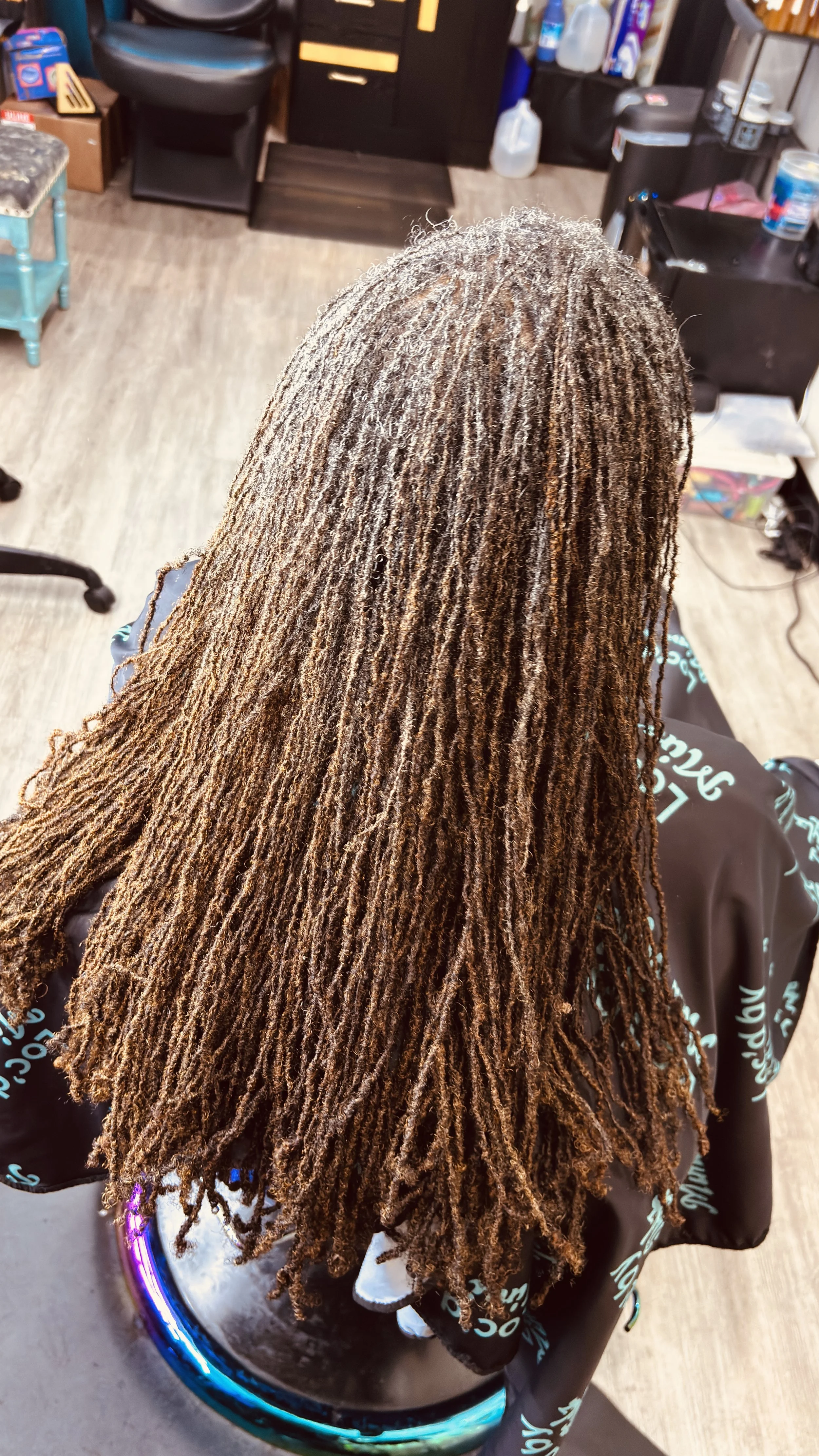 A person with long, textured dreadlocks hairstyle sitting in a salon chair, focusing on the hairstyle from the back. The salon has various equipment, products, and furniture visible in the background.