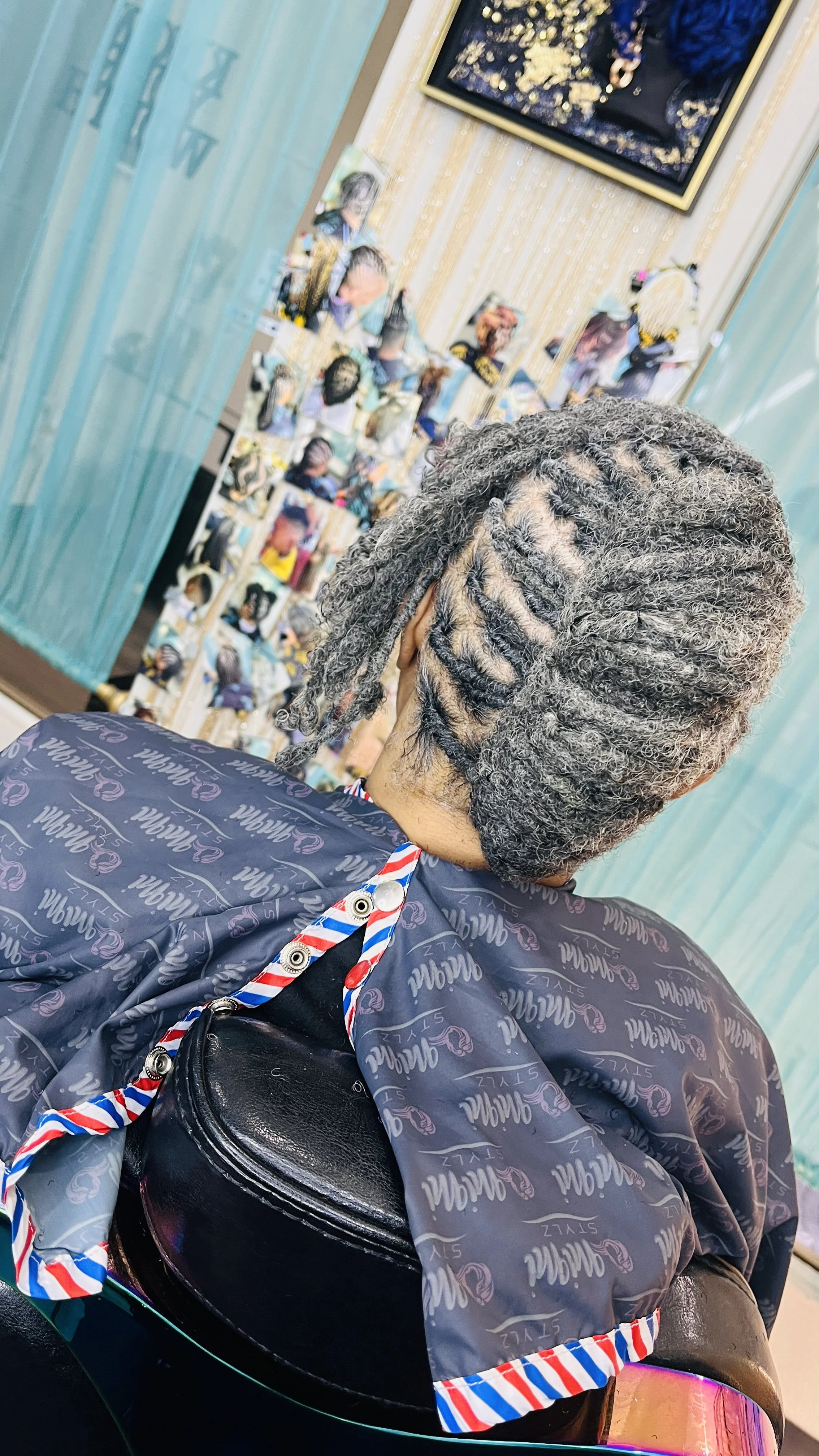 Back of a person with gray and black dreadlocks sitting on a black chair in front of a mirror with photos and artwork on the wall.