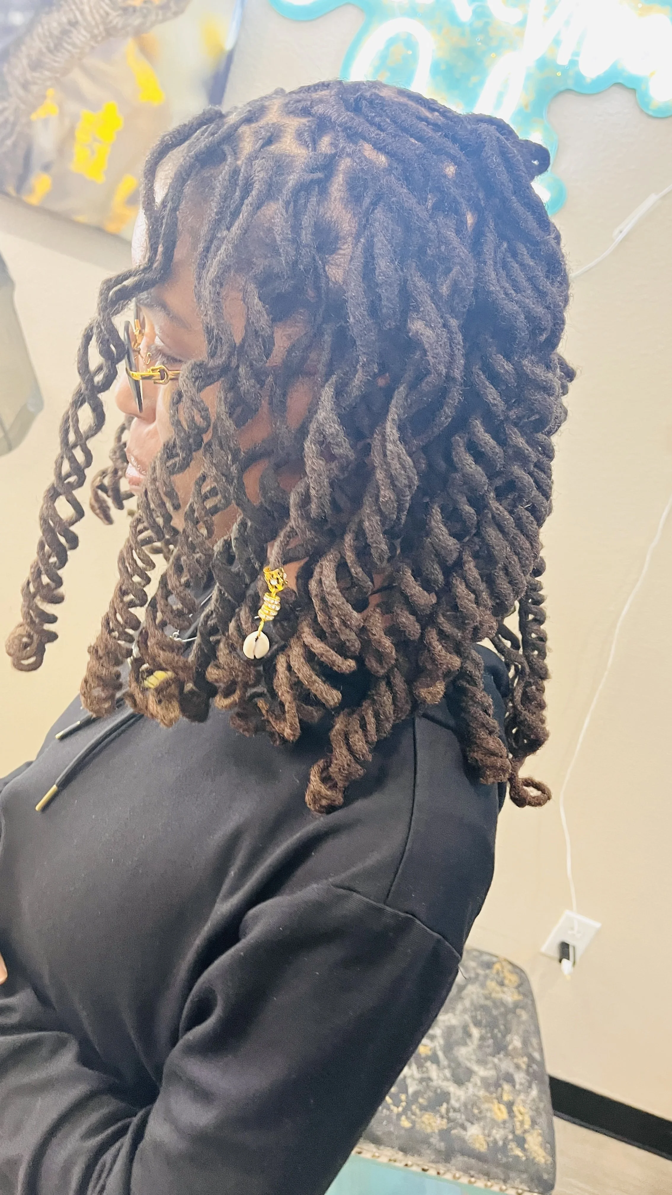 Side view of a woman with long locs styled in thick twists, wearing glasses with gold accents, dressed in a black hoodie, sitting on a chair with a patterned seat. A colorful wall art is visible in the background.