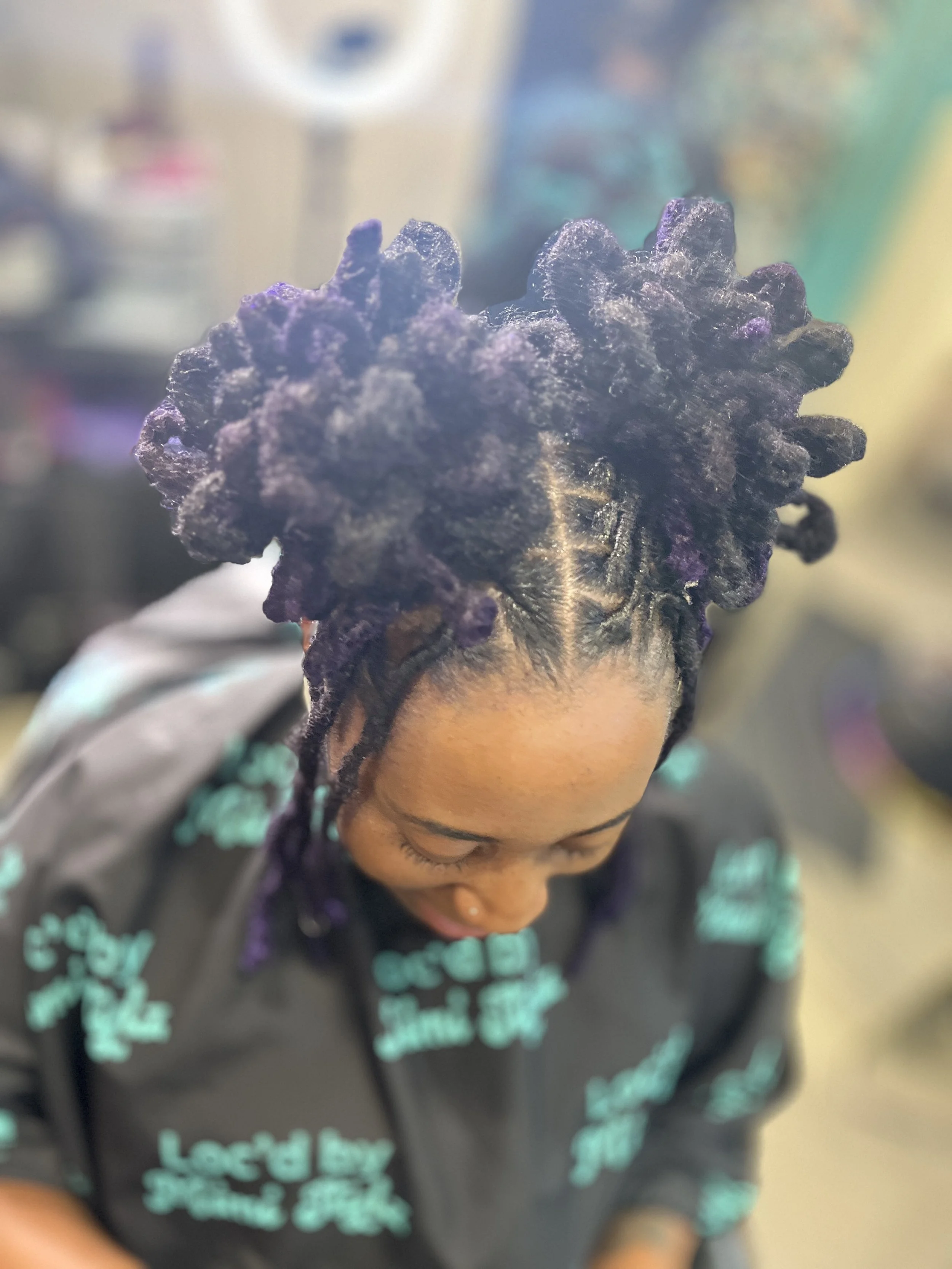 A person with long braided hair styled into large, textured purple locs, looking down, wearing a dark hoodie with turquoise text.