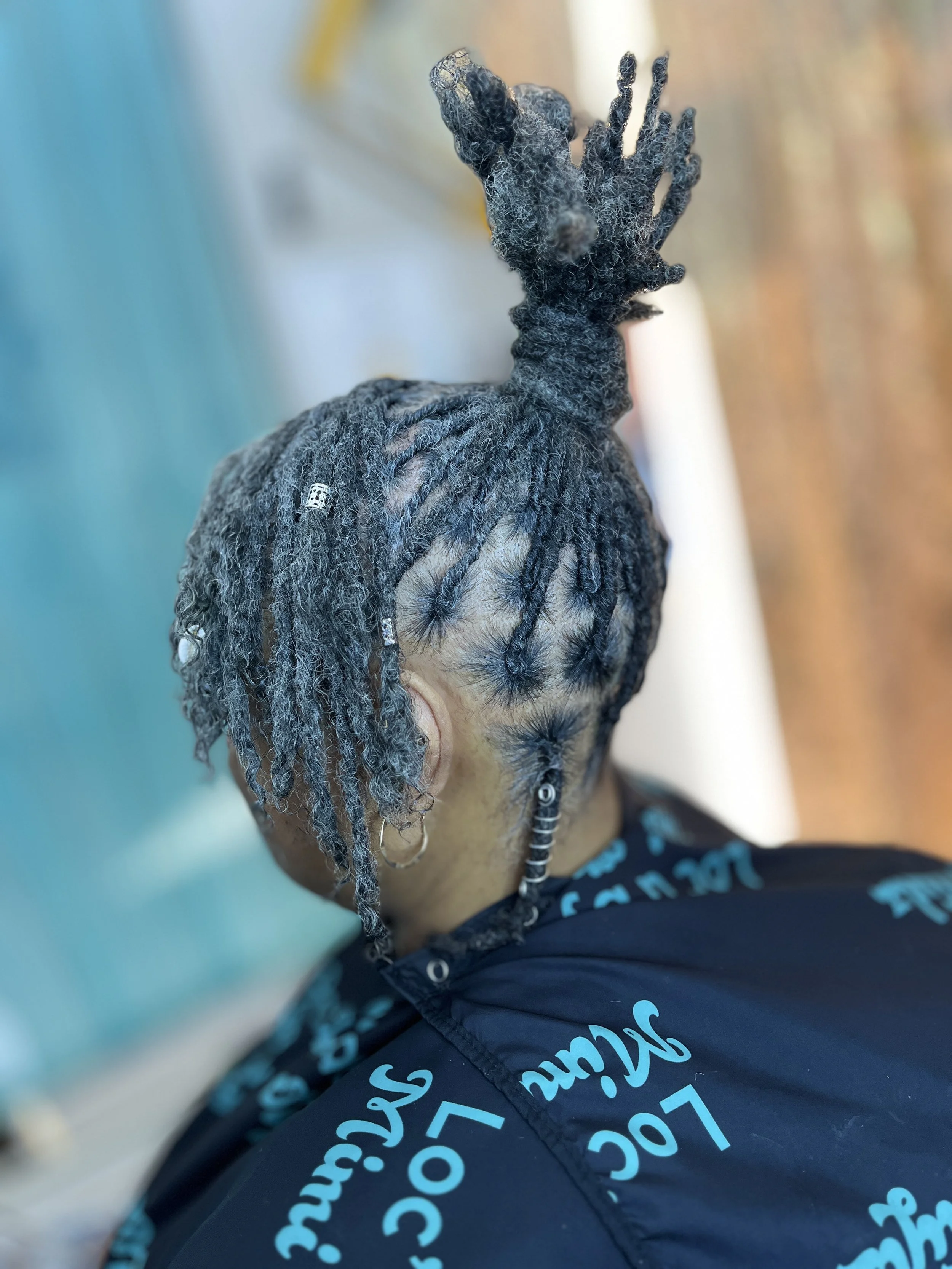 Close-up view of a person's head with dreadlocks, partial view of their face, wearing earrings, and a dark patterned shirt, with a blurred background.