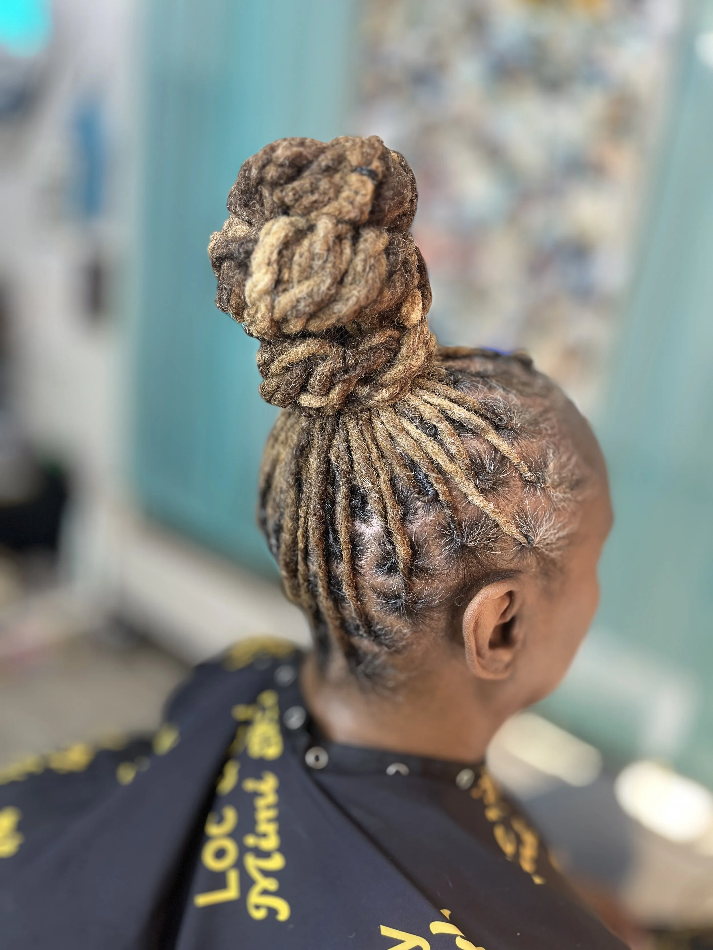 Close-up of a person's head with styled and twisted blonde dreadlocks, seen from the back. The person is wearing a black shirt with yellow lettering.