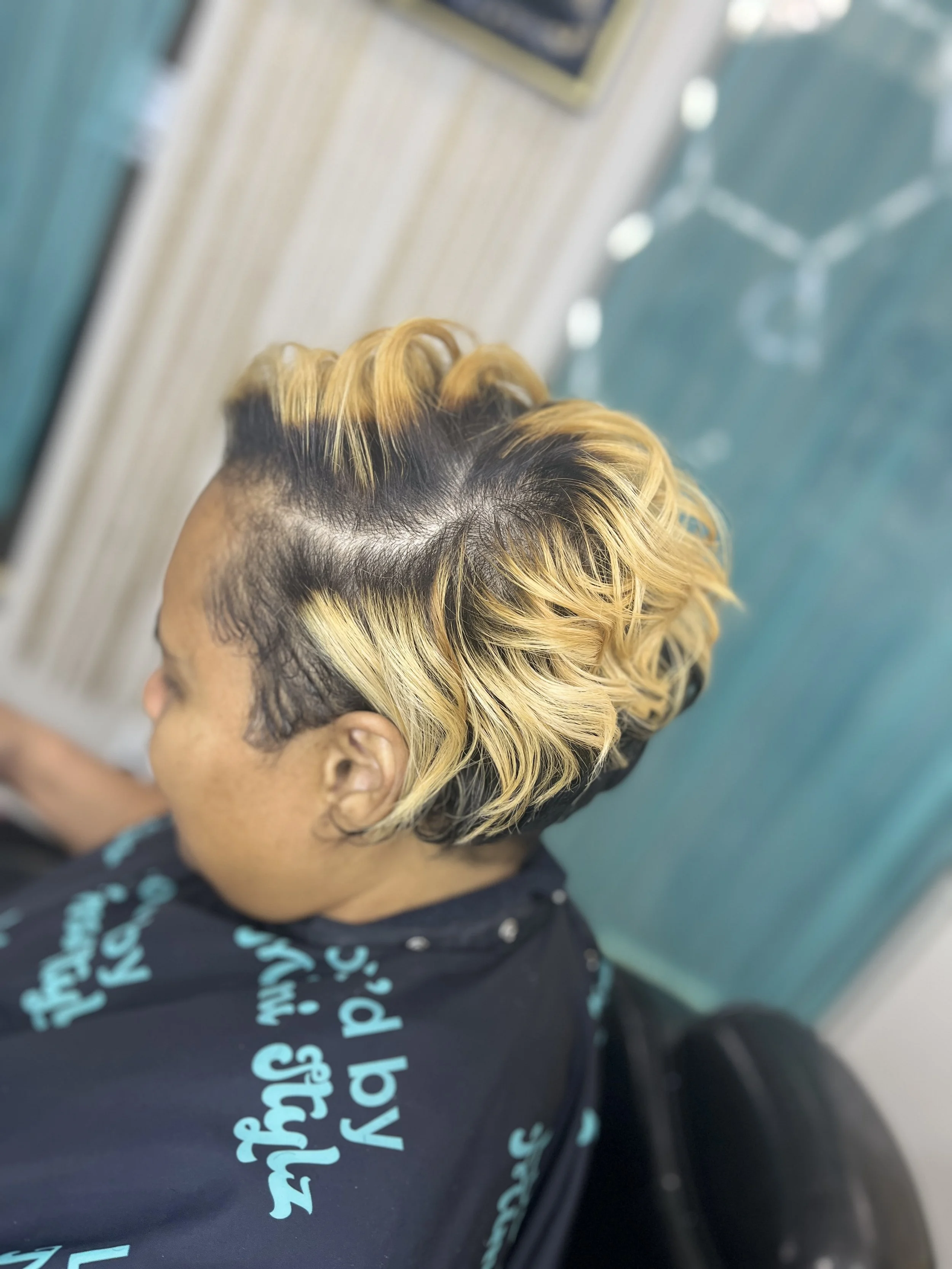 Close-up of a person's head with a trendy hairstyle featuring short sides and longer, wavy, blonde-tinted hair on top, wearing a black shirt with blue text.