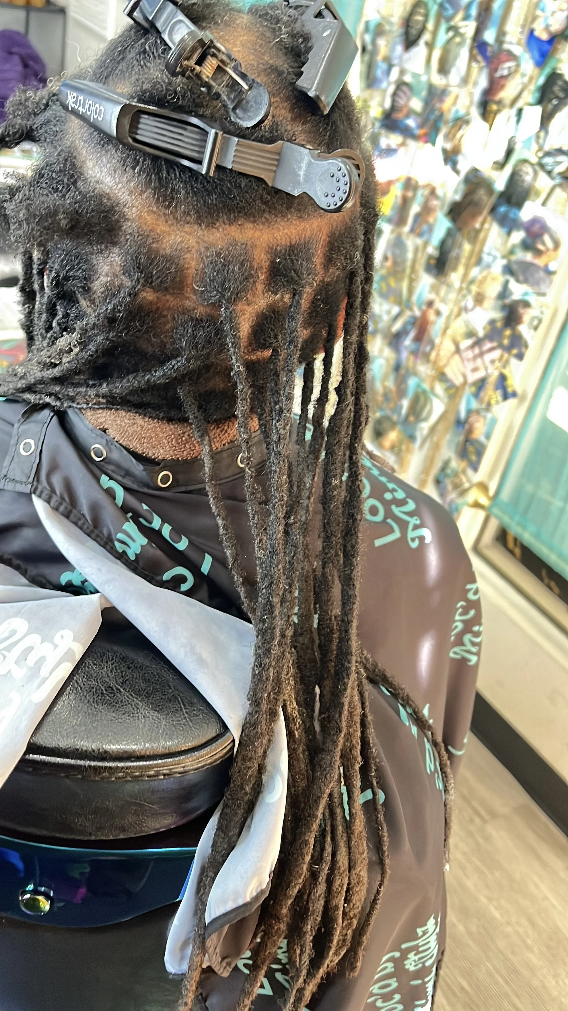 Person with long dreadlocks in a salon chair, facing away, with a hair clip and scissors on top, and a shop with colorful posters or pictures in the background.