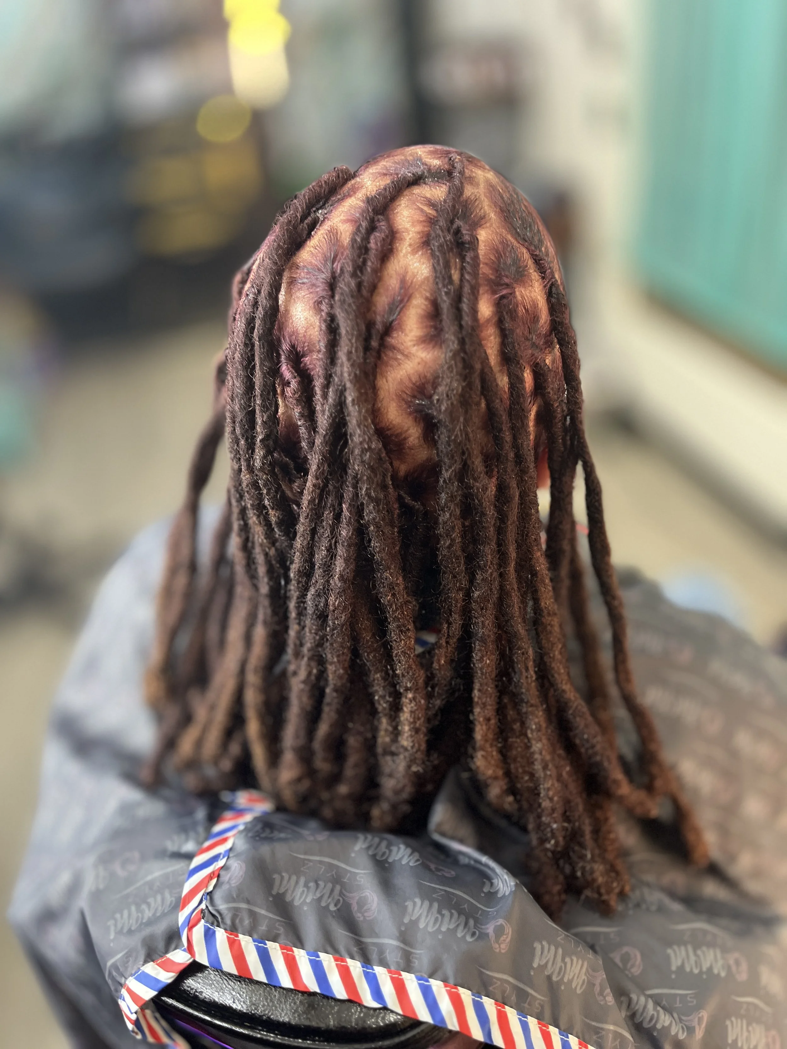 The image shows a person with long dreadlocks viewed from behind, sitting in a space with blurred background, with a wrapped gift box in the foreground.