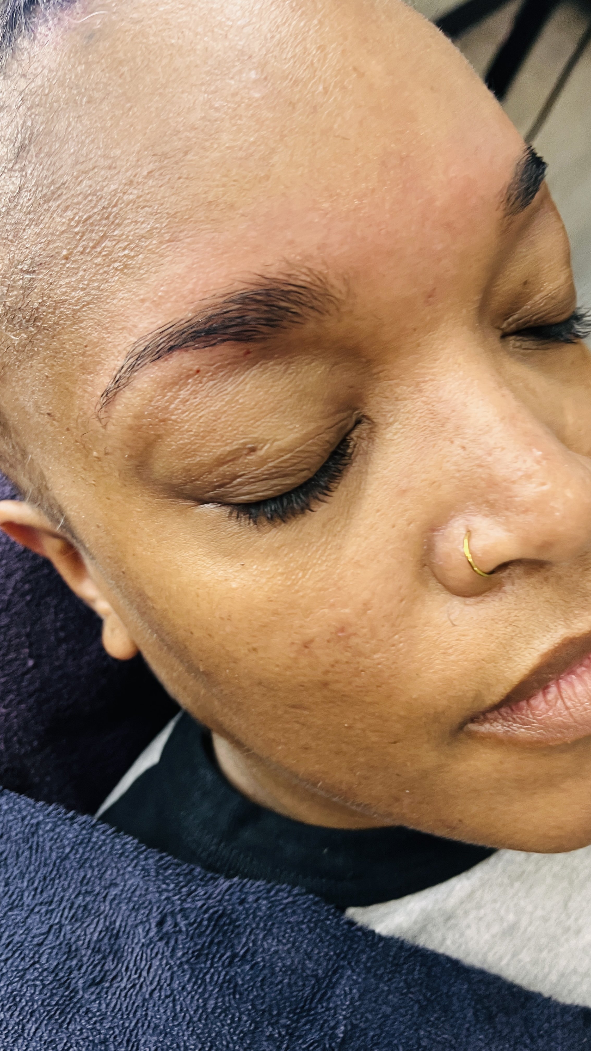 Close-up of a woman's face with closed eyes, showing well-groomed eyebrows, long eyelashes, and a nose ring. The skin is smooth with a natural complexion.