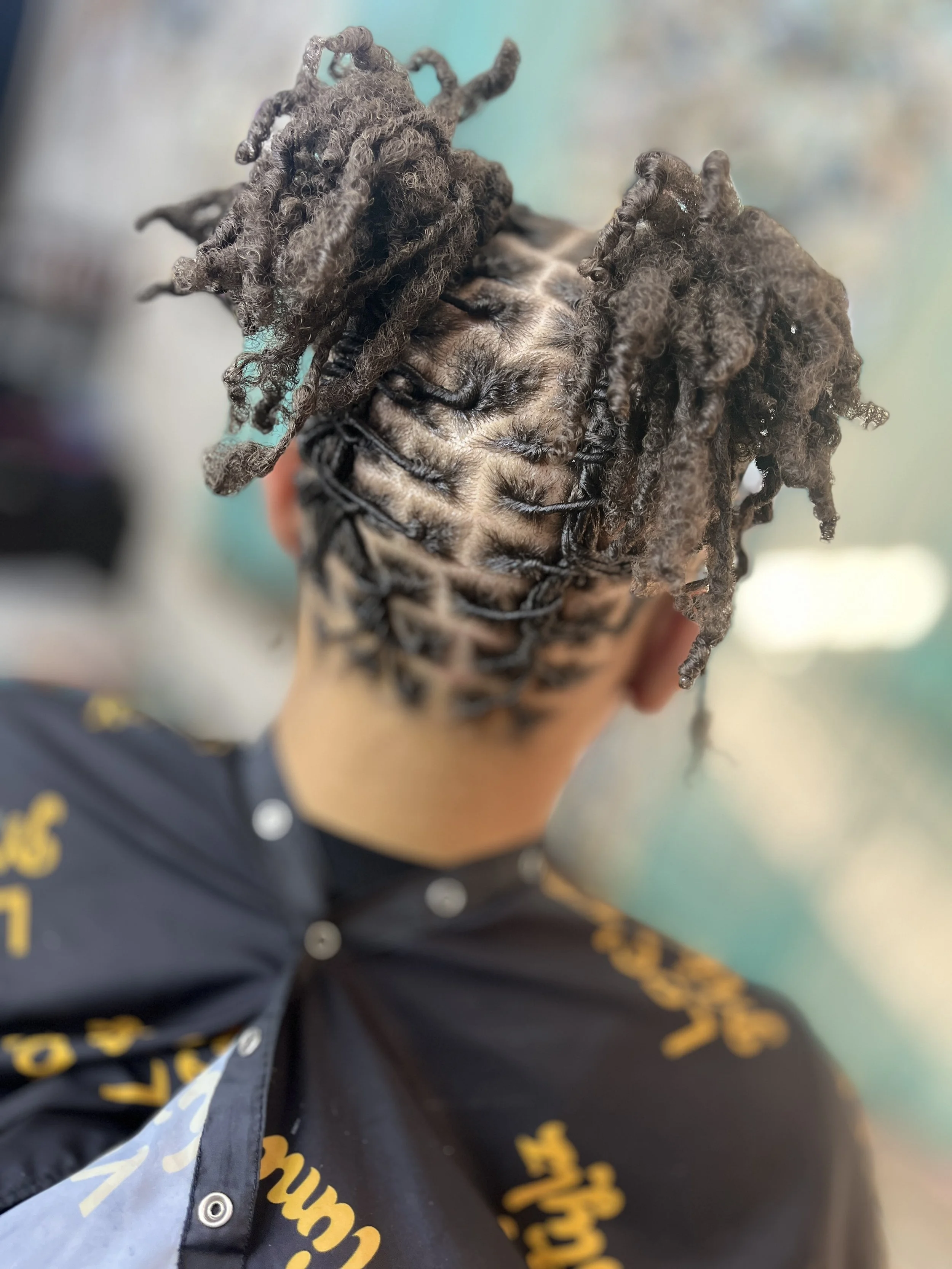 Back of a person's head with styled dreadlocks hair, wearing a black shirt with yellow lettering.