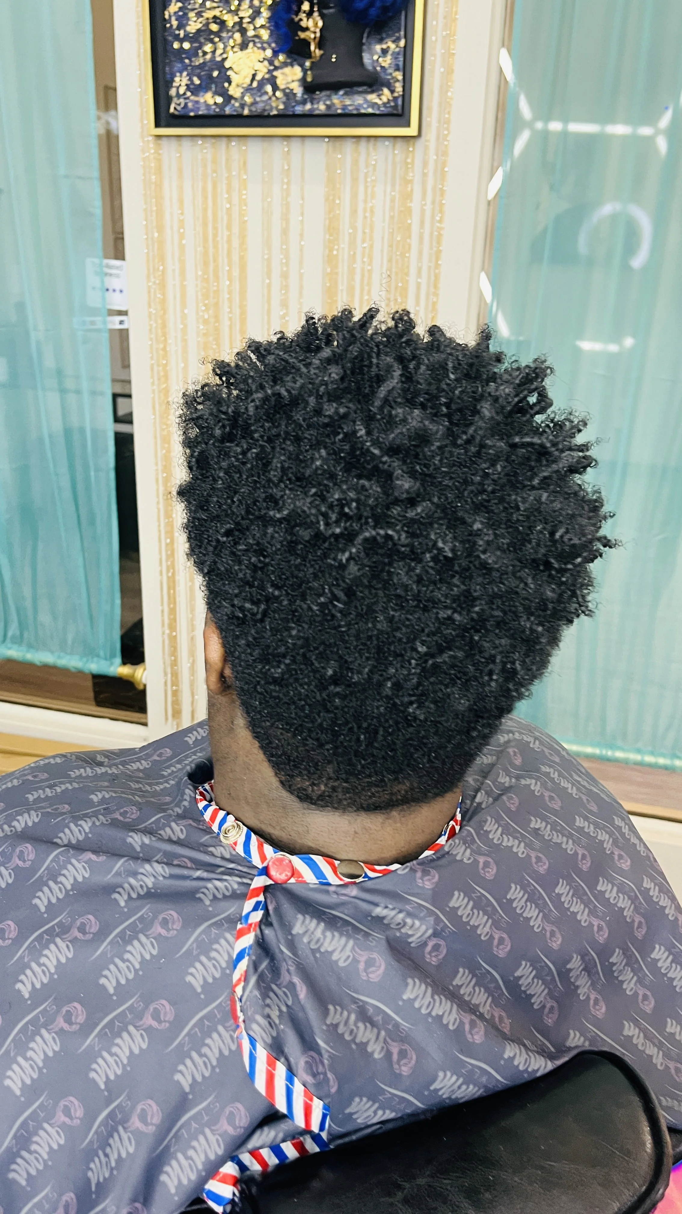Back view of a person with short, curly black hair at a salon, wearing a cape with a patterned design, sitting in a chair.