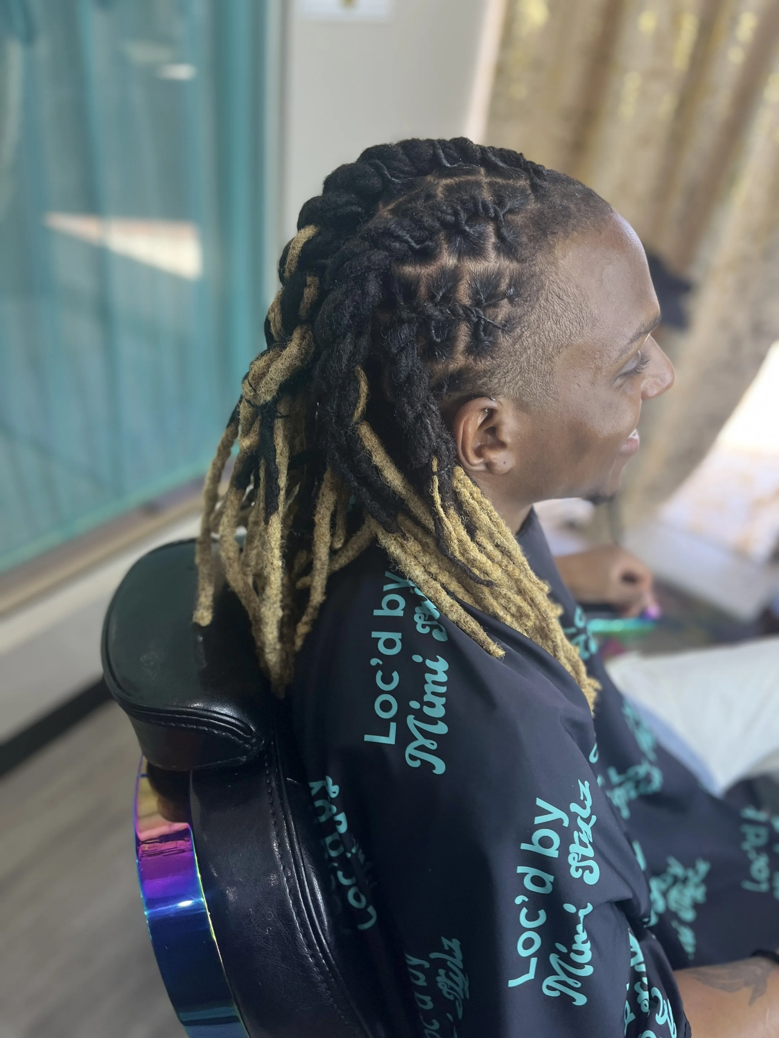 Profile of a person with styled dreadlocks, shaved sides, sitting on a black chair, wearing a black shirt with light blue text, indoor setting with a window and bathroom tiles visible.