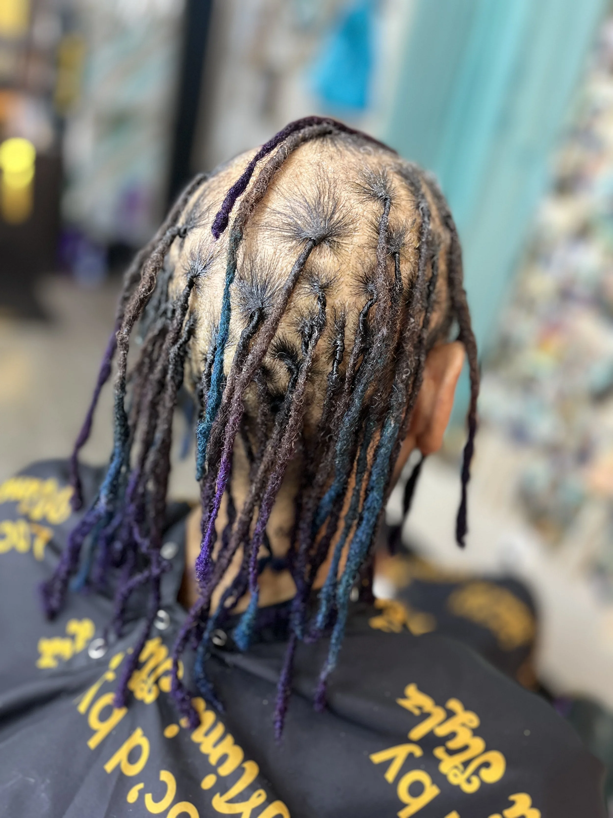 View of a person's head from behind with long dreadlocks featuring strands dyed in purple, blue, and brown. The background appears to be a store or salon with shelves and colorful items. The person is wearing a black shirt with yellow lettering.