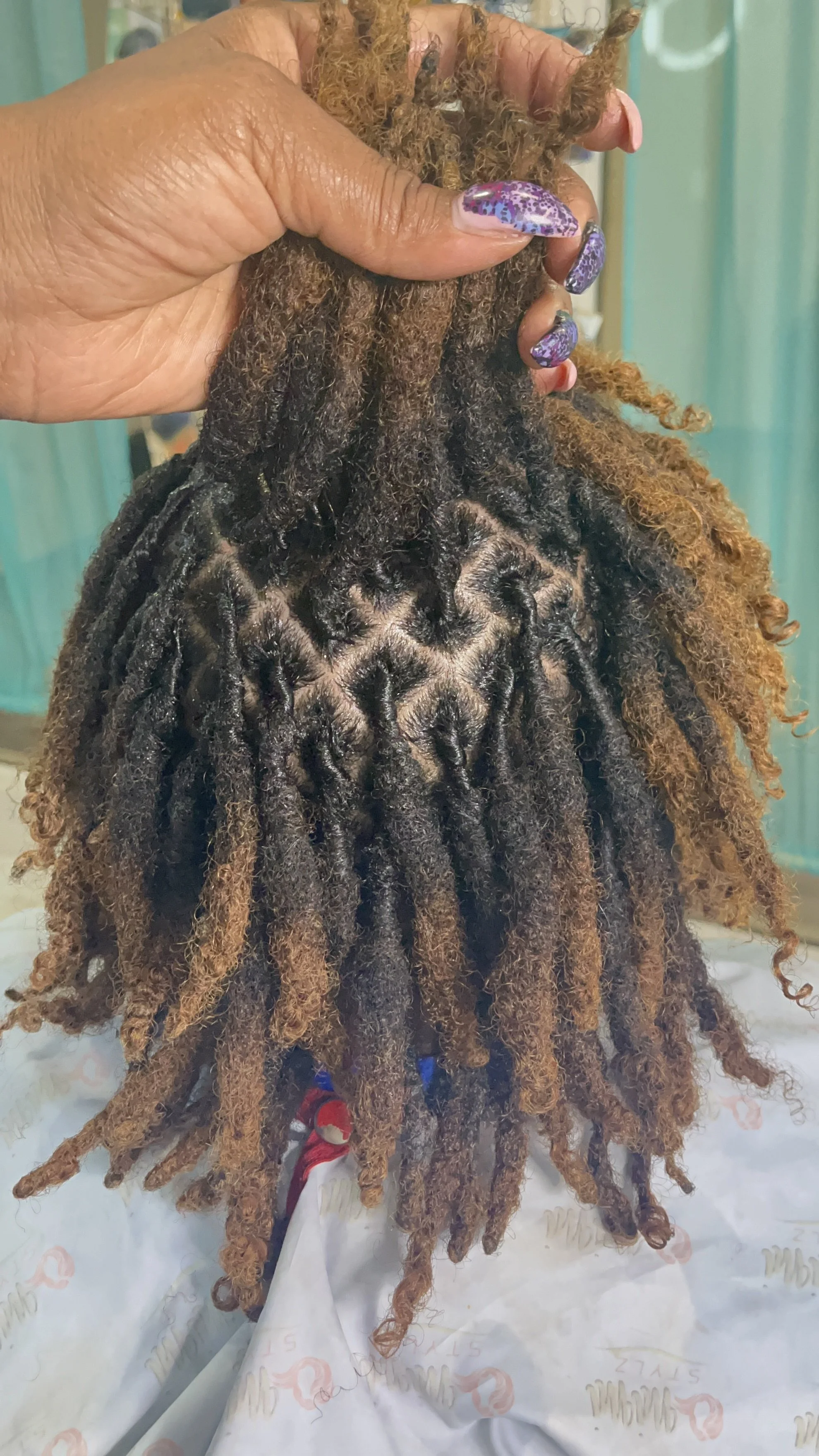 A person with intricately styled dreadlocks held up by their hand, showing a gradient of dark to light brown and blonde colors, with purple and blue floral decorated nails.