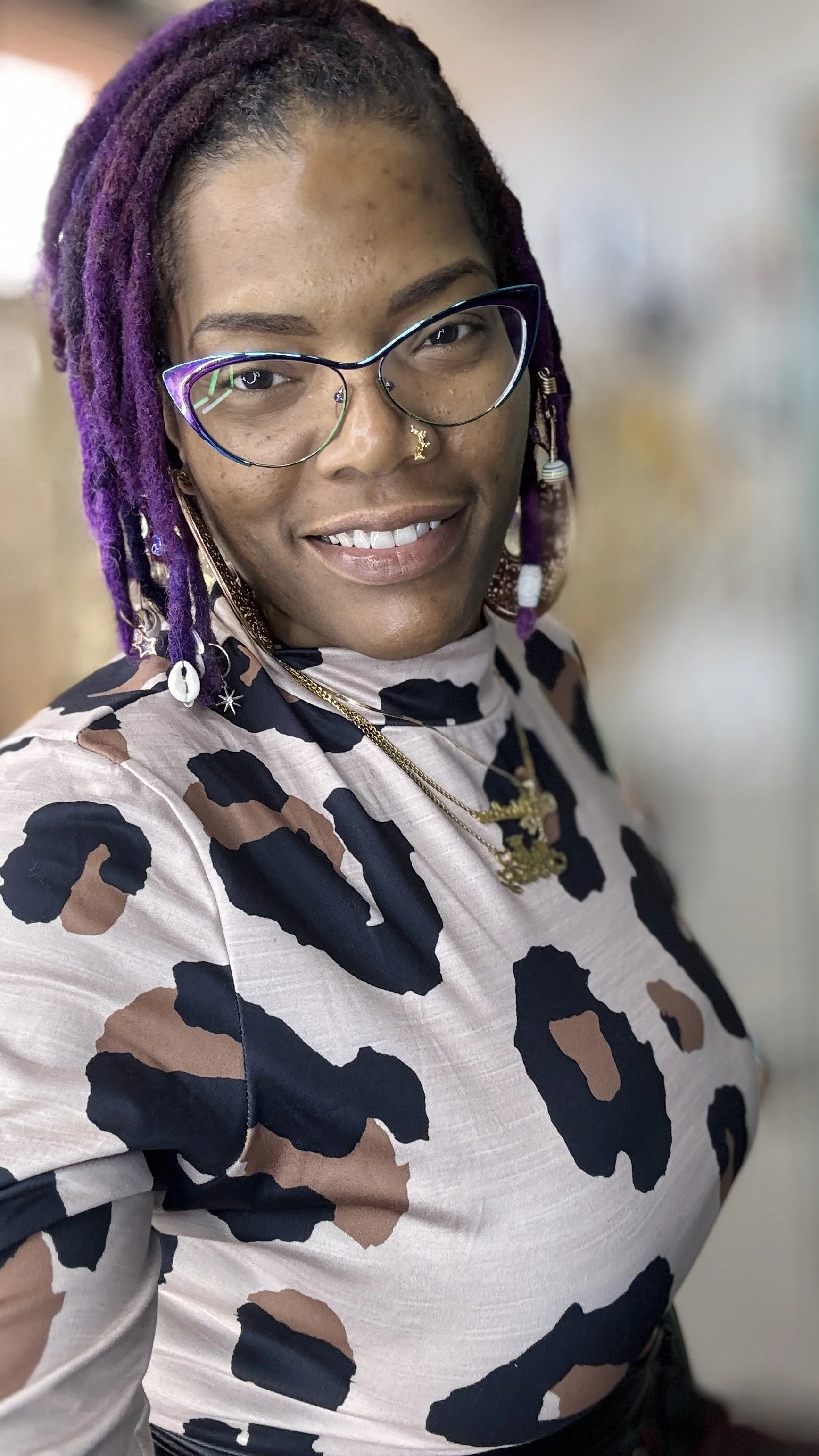 A woman with purple dreadlocks, wearing glasses and a leopard print top, is smiling at the camera.