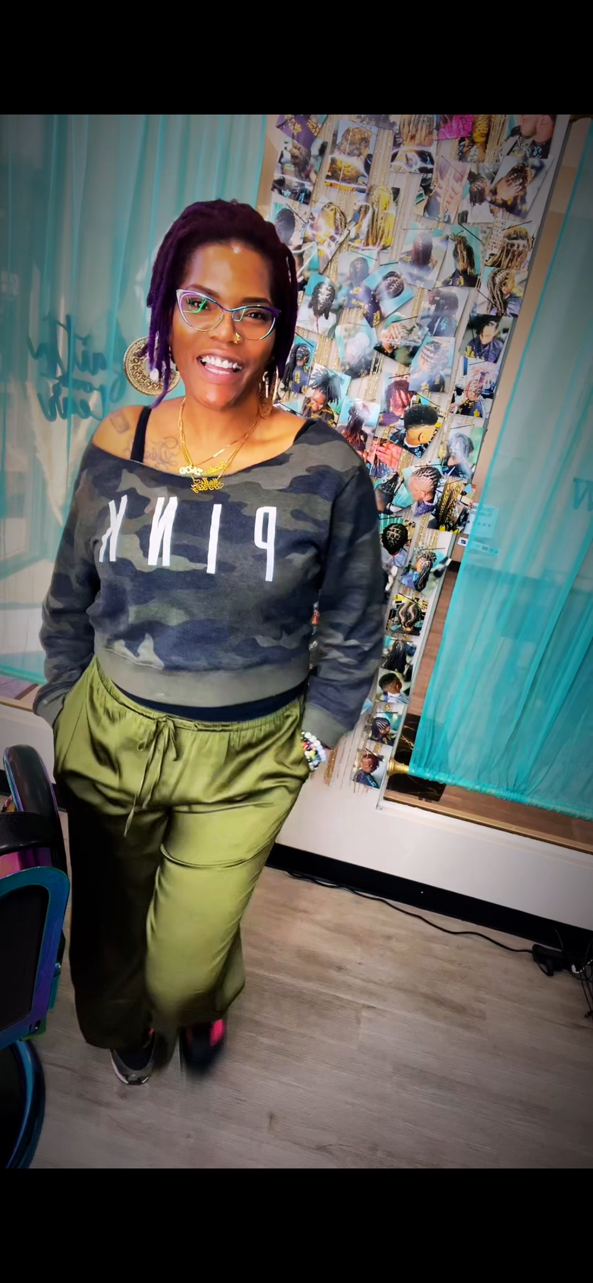 Women standing in front of a collage of photos, wearing glasses, a camo sweatshirt, and green pants.