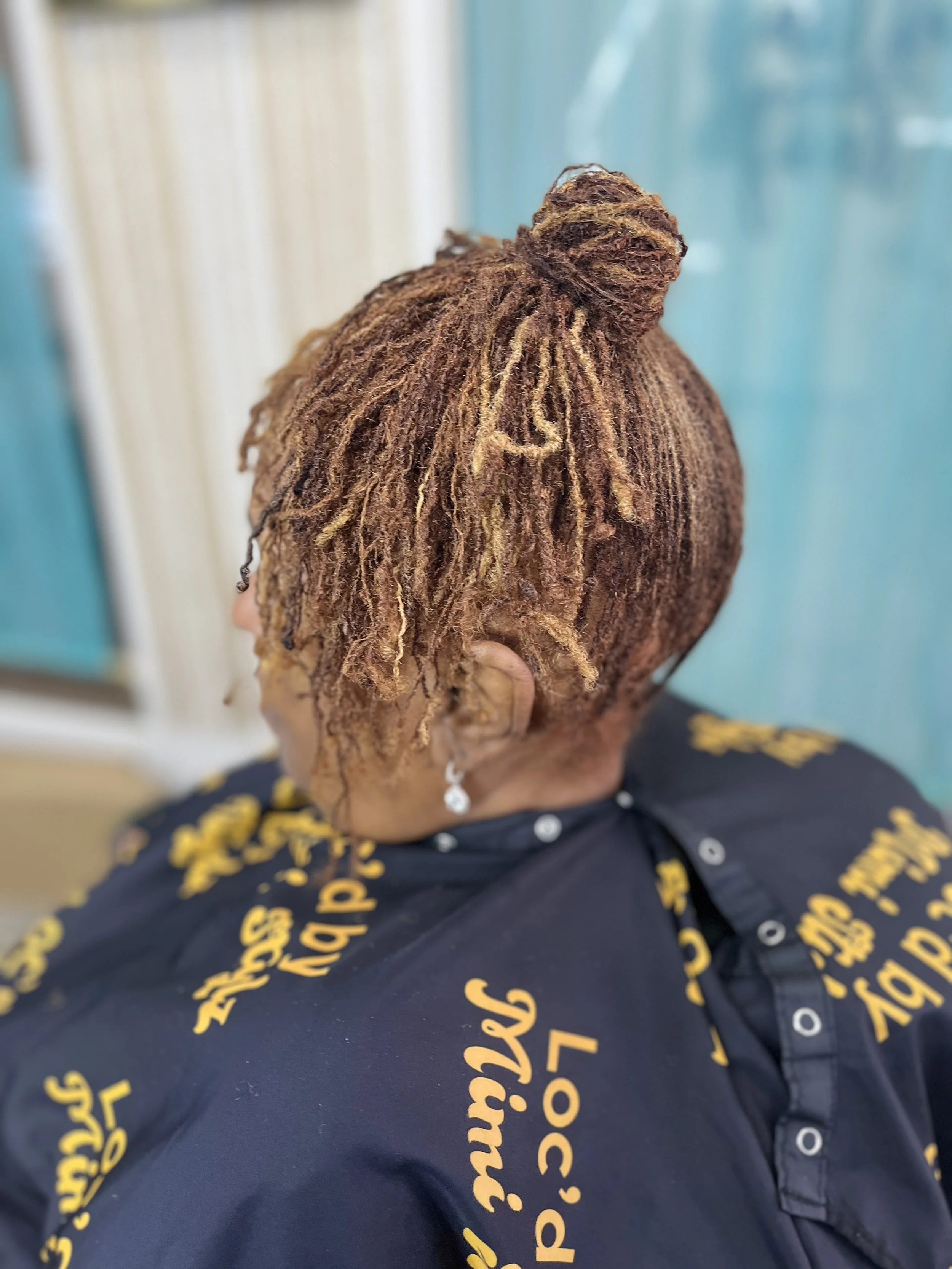 A person with styled, brown dreadlocks with blonde highlights, wearing a black shirt with yellow lettering, holding their head with one hand.