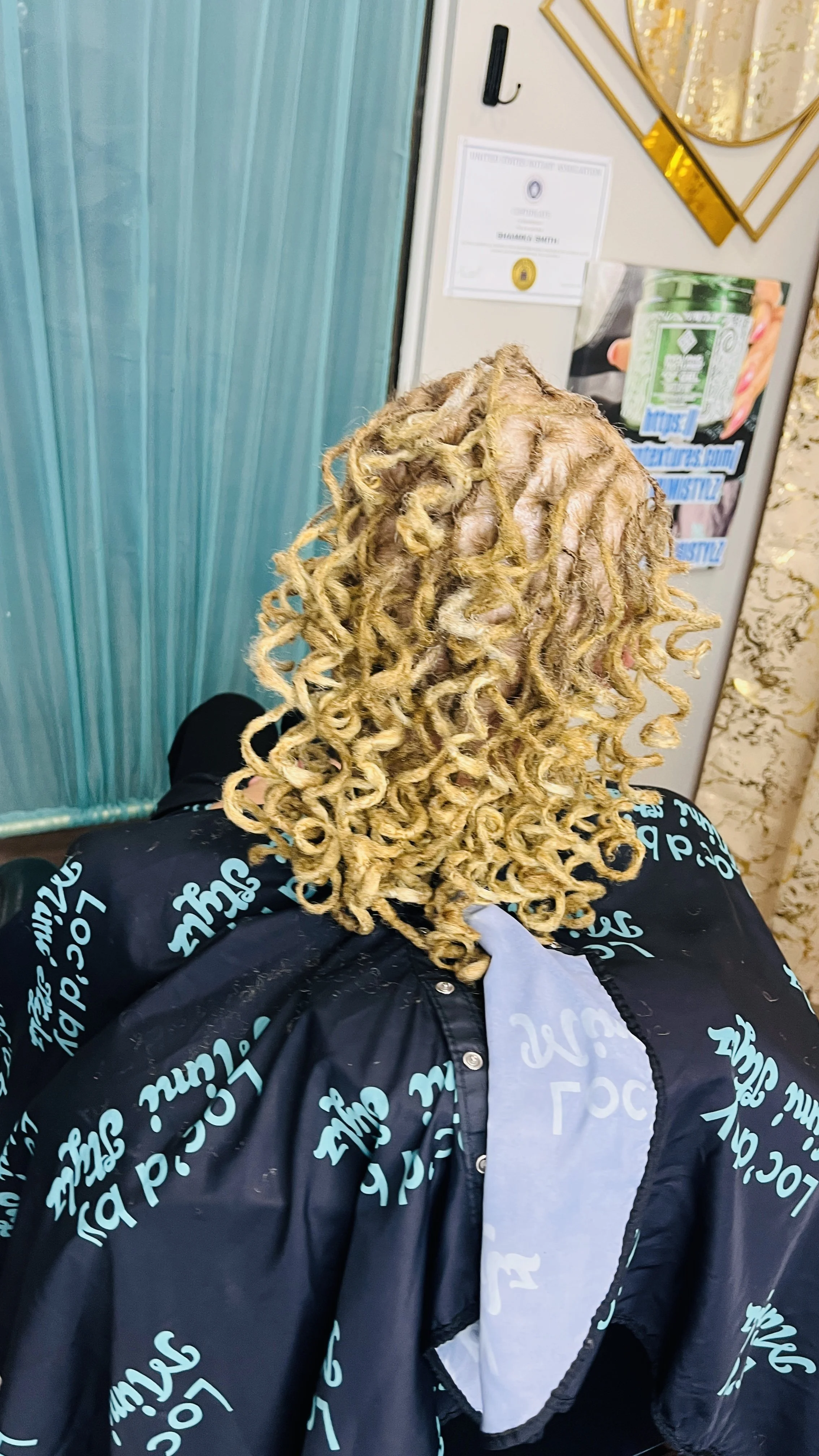 Person with long, curly blonde hair sitting in a waiting area, wearing a black jacket with printed turquoise text.