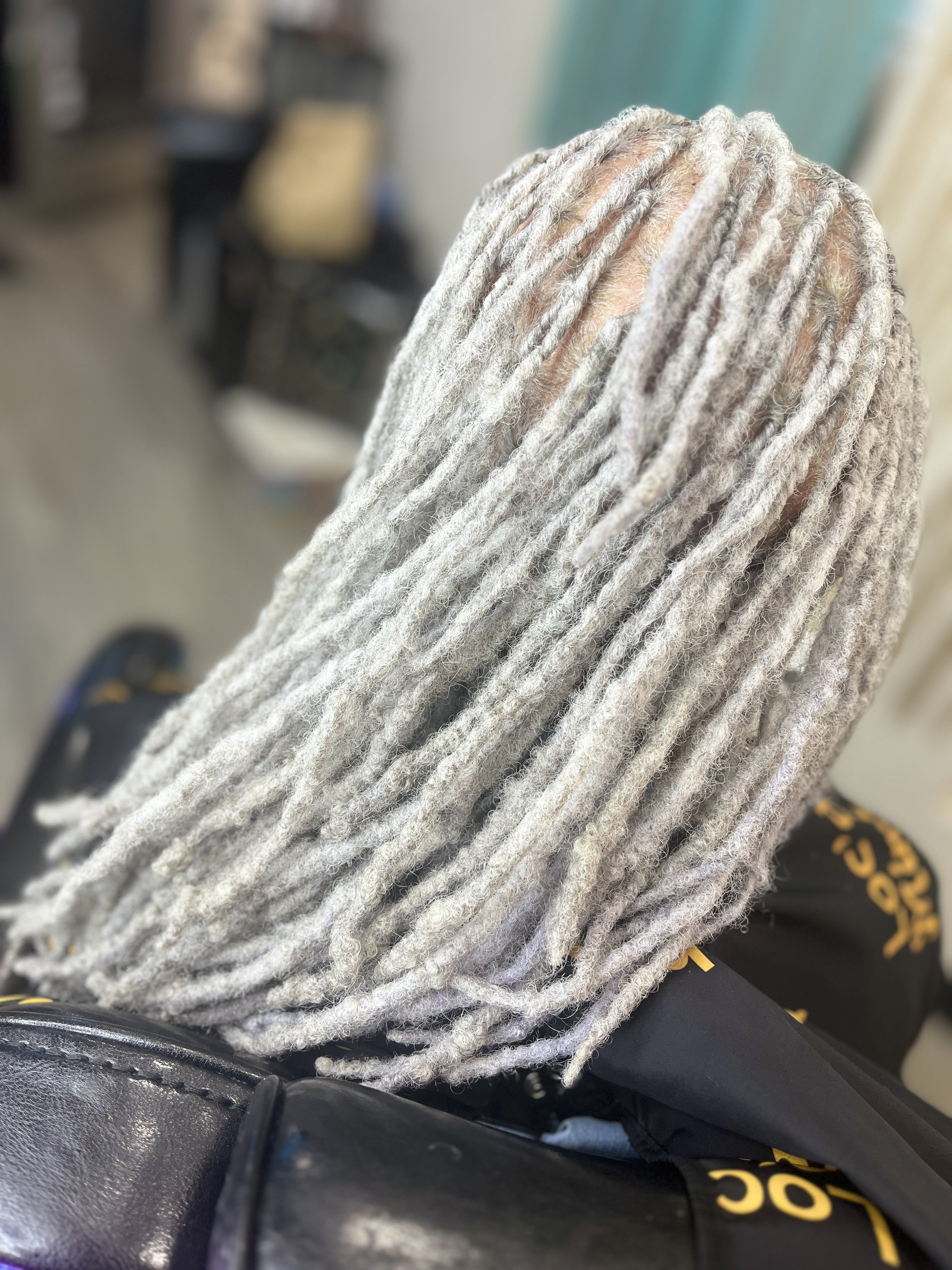 Close-up of a person's dreadlocks with a grayish tone, wearing a gray knit hoodie or sweater, in an indoor setting with blurred background.