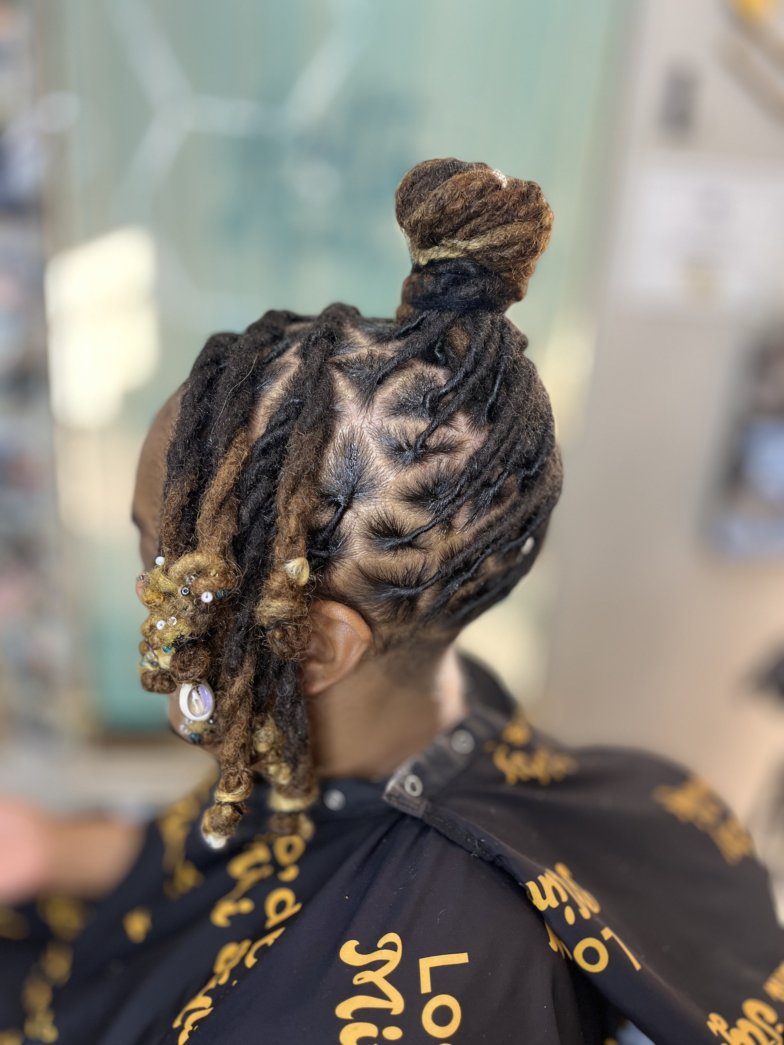 Close-up of a woman with intricately styled dreadlocks with beads and accessories, wearing a dark shirt with yellow text.