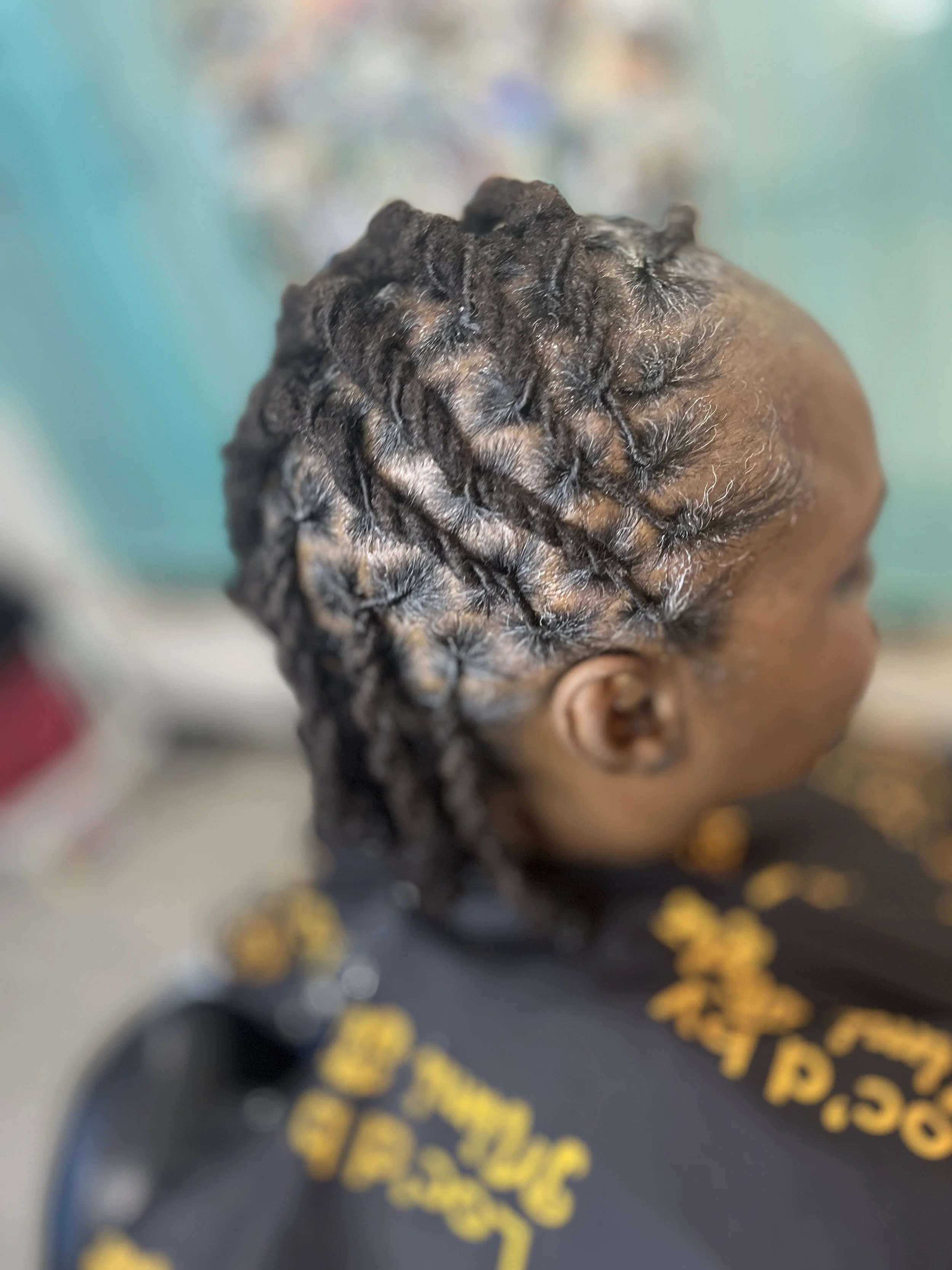 Close-up of a person with braided hair. They are wearing a black shirt with yellow text. The background is blurred.