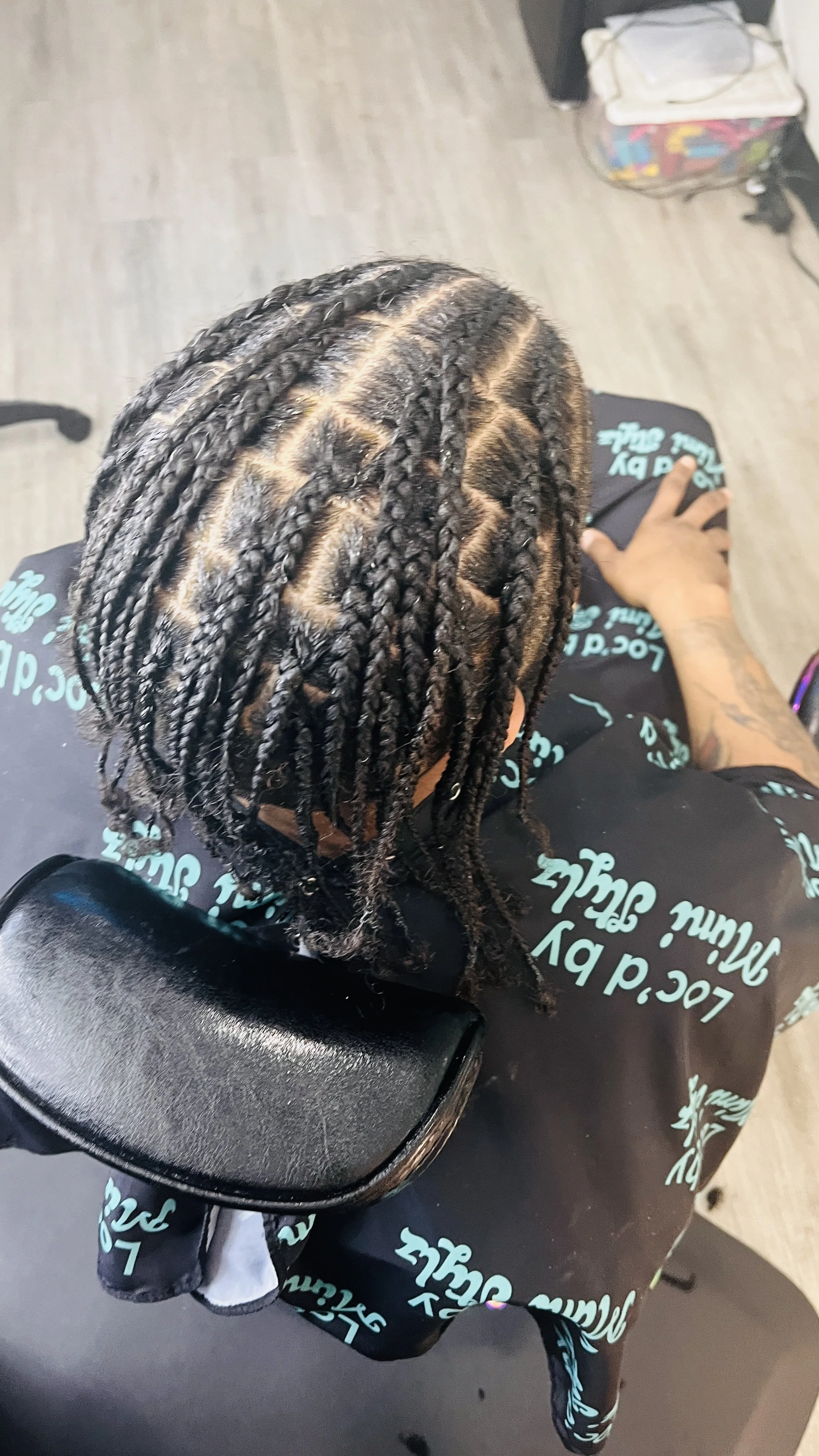 View from above of a person with freshly braided hair with a middle parting, sitting in a chair in a salon. The person is wearing dark clothing with light blue text, and there is a black bag or purse on their lap. The salon has a wooden floor, with s