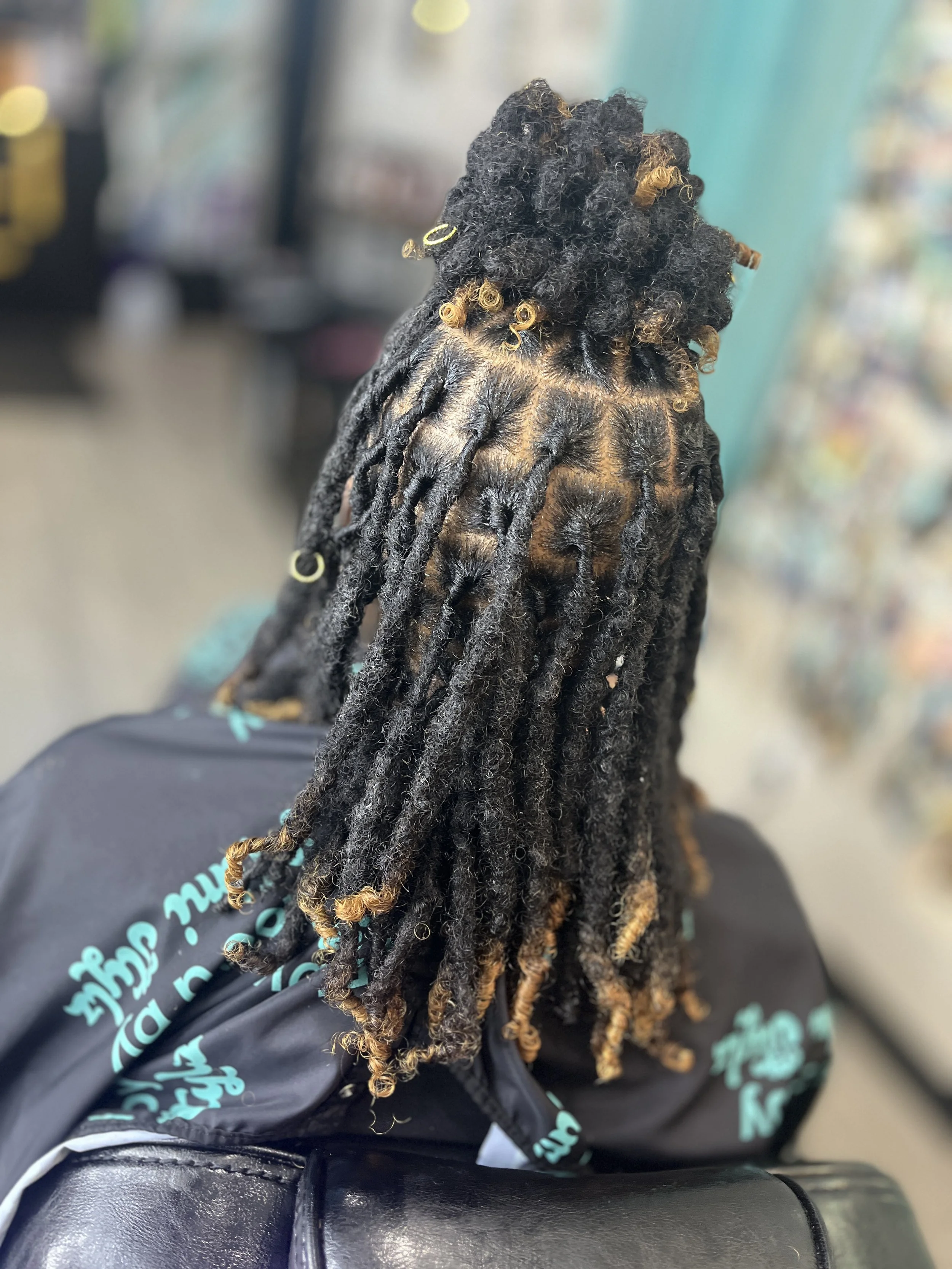 Close-up image of a person's head with long, thick dreadlocks with gold rings, showing black and brown hair. The background is blurred, and part of the person's clothing with writing is visible.