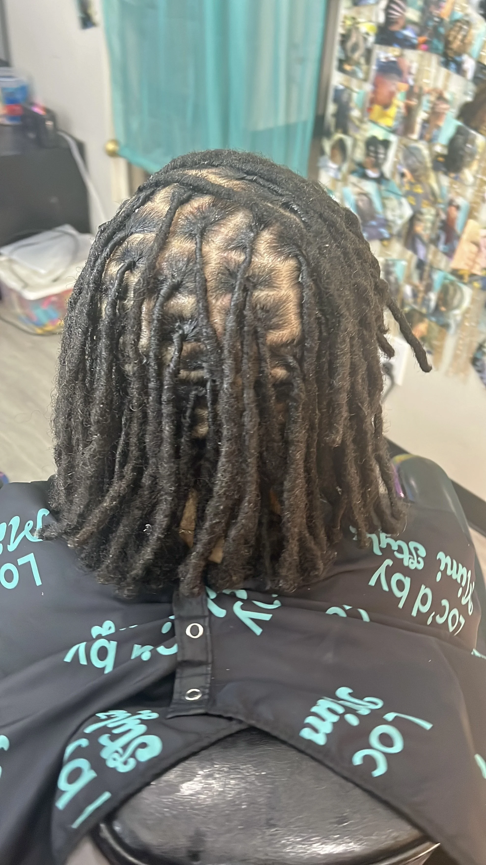 A person with short dreadlocks sitting in a salon chair, with their back to the camera. The background includes a wall of photos, a turquoise curtain, and salon-related items.
