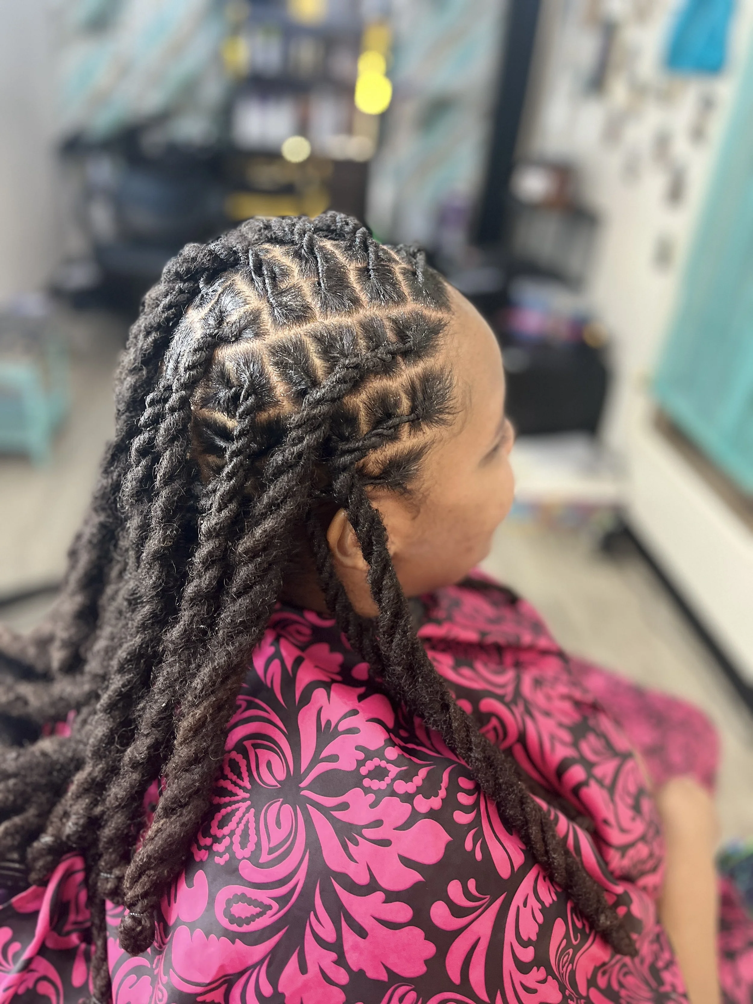 Close-up of a person with styled, twisted dreadlocks, wearing a pink and purple patterned dress in a salon.