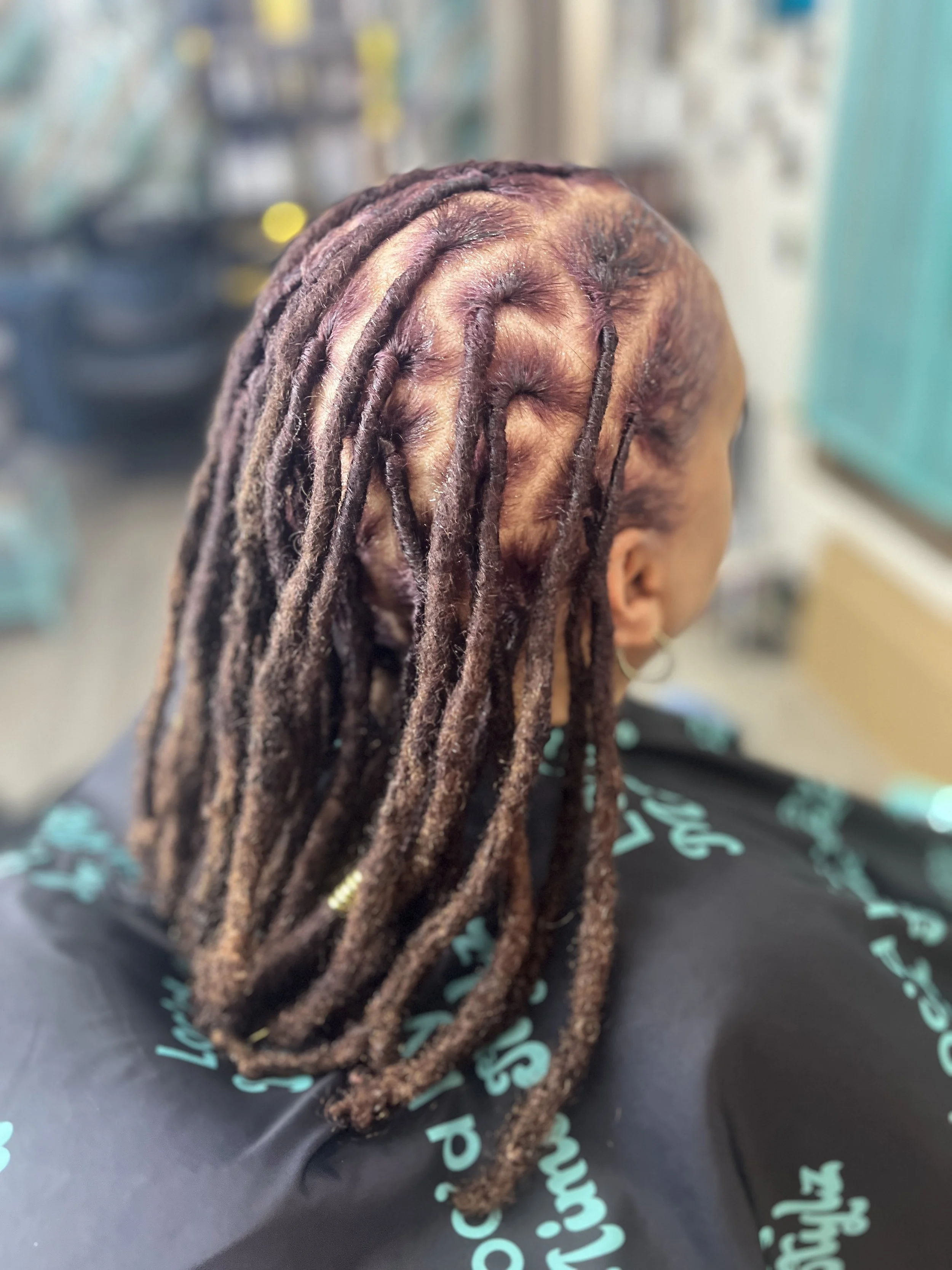 Close-up of a person's head with neatly styled dreadlocks, sitting on a black surface with teal script writing, with a blurred store background.